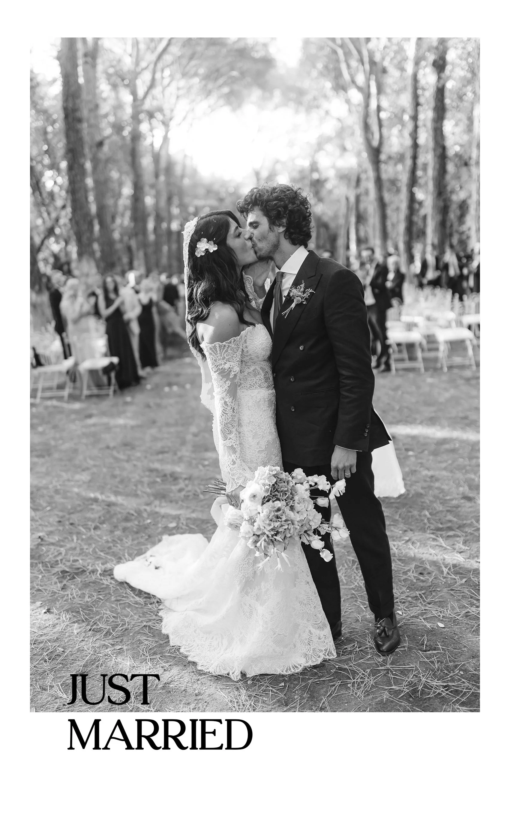 A newlywed couple in wedding attire sharing a kiss outdoors during their wedding ceremony, with guests in the background and holding a bouquet of flowers.