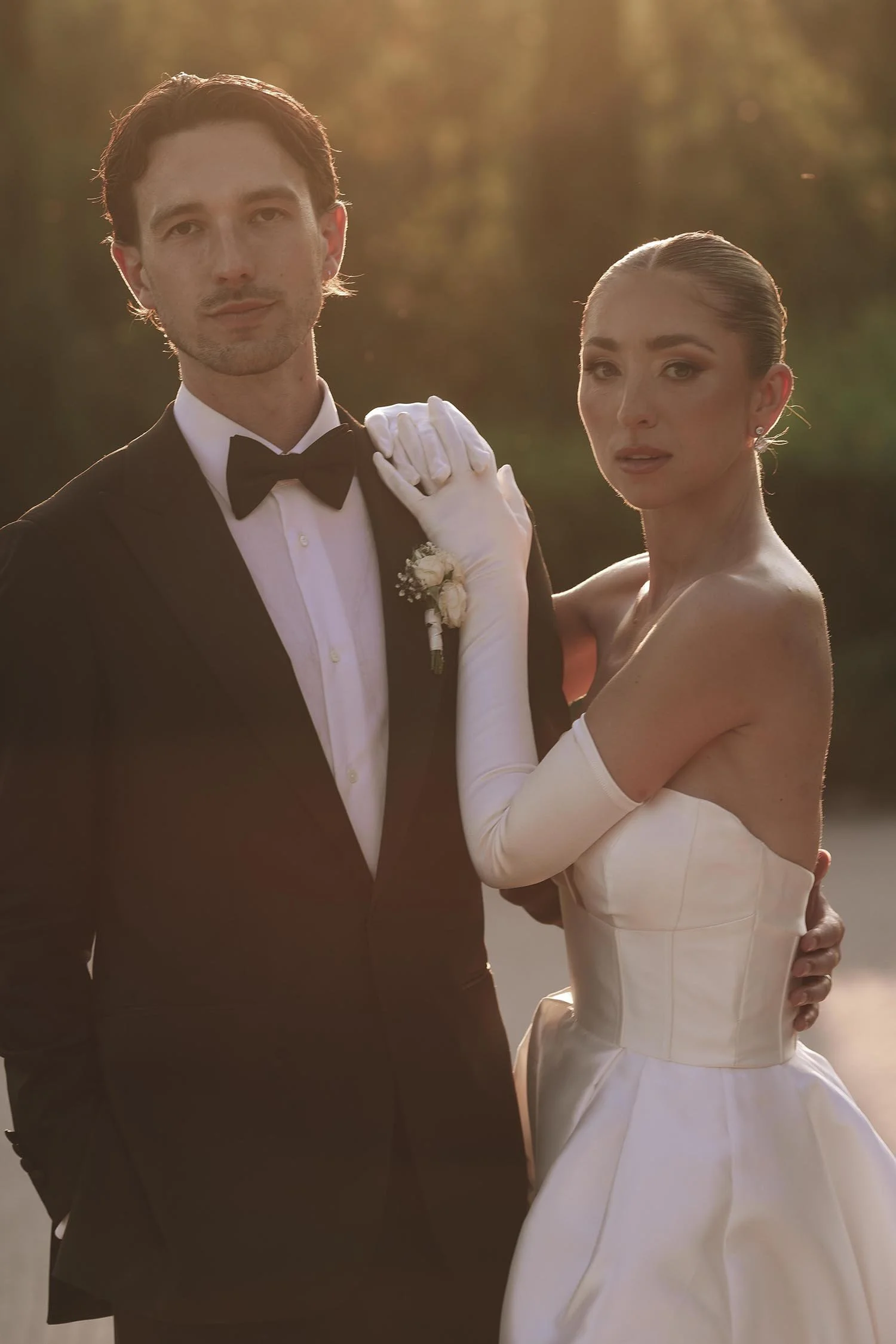 Bride and groom standing together and looking in camera in the driveway between the cypresses with the sun setting in the background