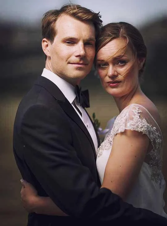 A couple dressed in wedding attire standing close outdoors, with the woman in a white gown and the man in a black tuxedo, both looking at the camera.