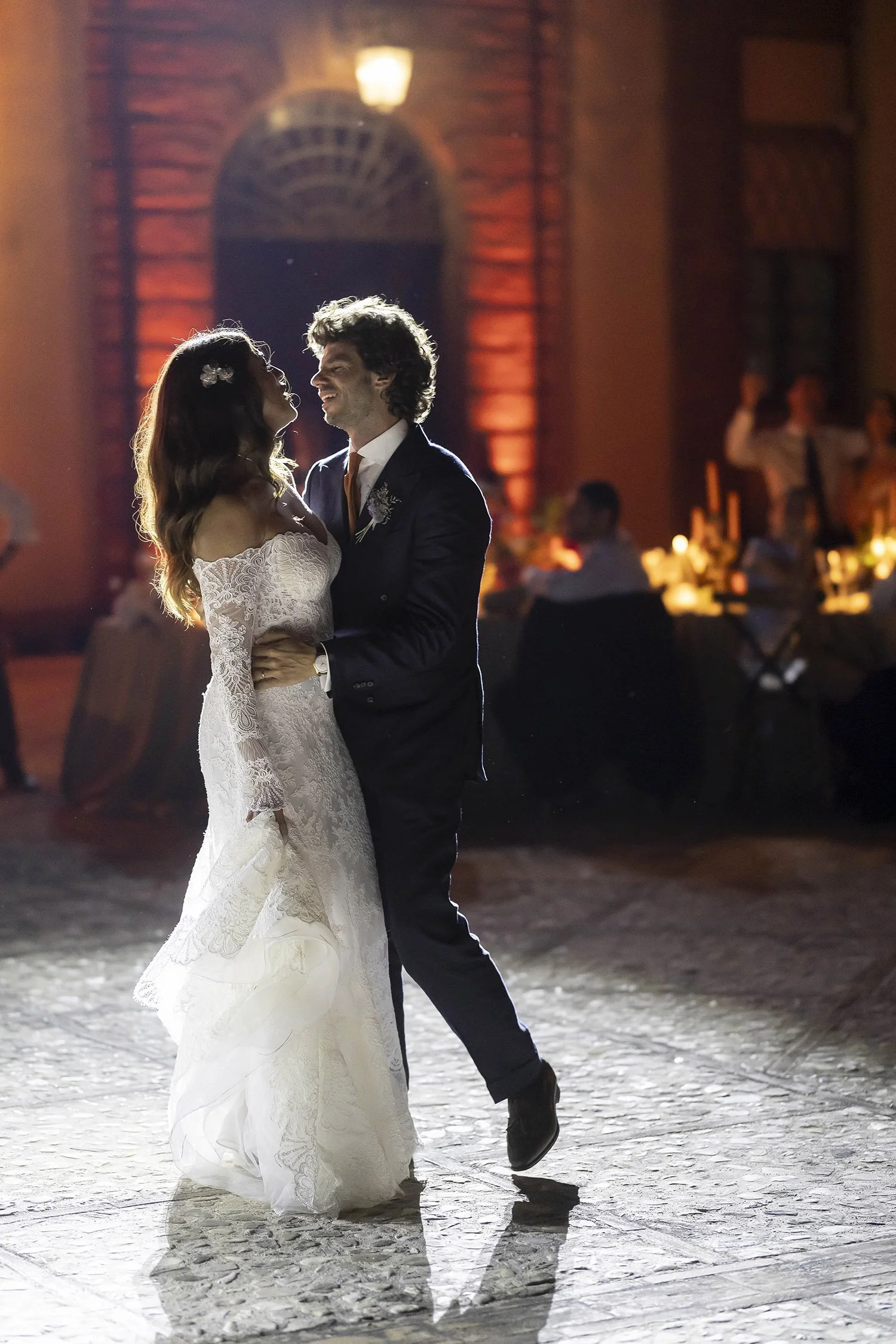 A bride and groom dancing at their wedding reception, with the bride in a lace wedding dress and the groom in a dark suit, in a warmly lit venue with seated guests in the background.