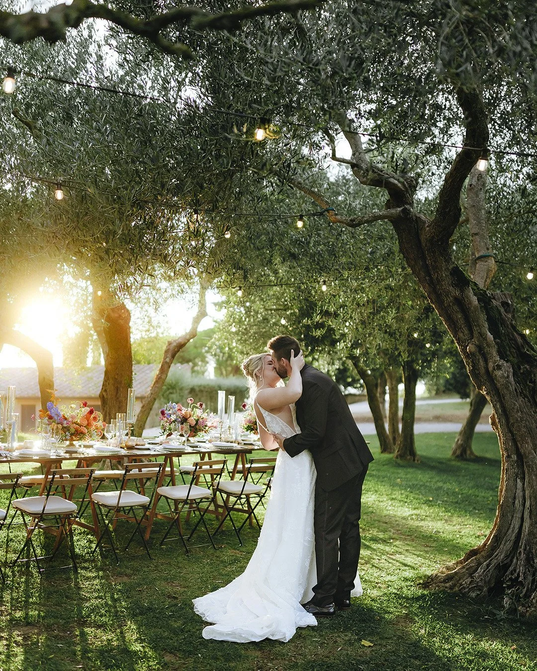 Bride and groom holding each other under an olive tree with the sun setting on the dinner table