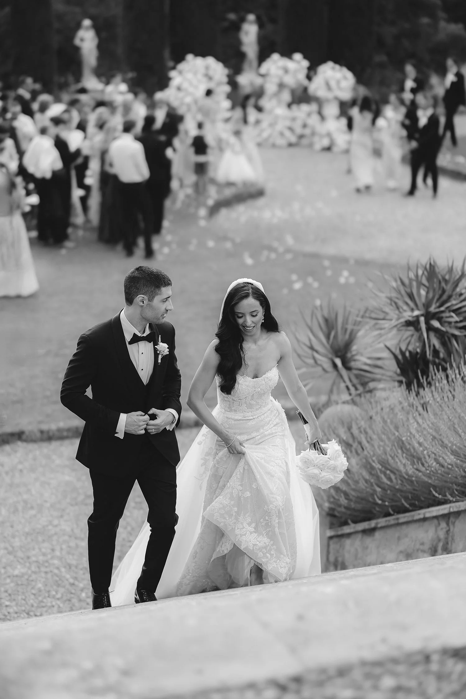 Black and white photo of a bride and groom walking outside at a wedding reception, with guests gathered in the background.