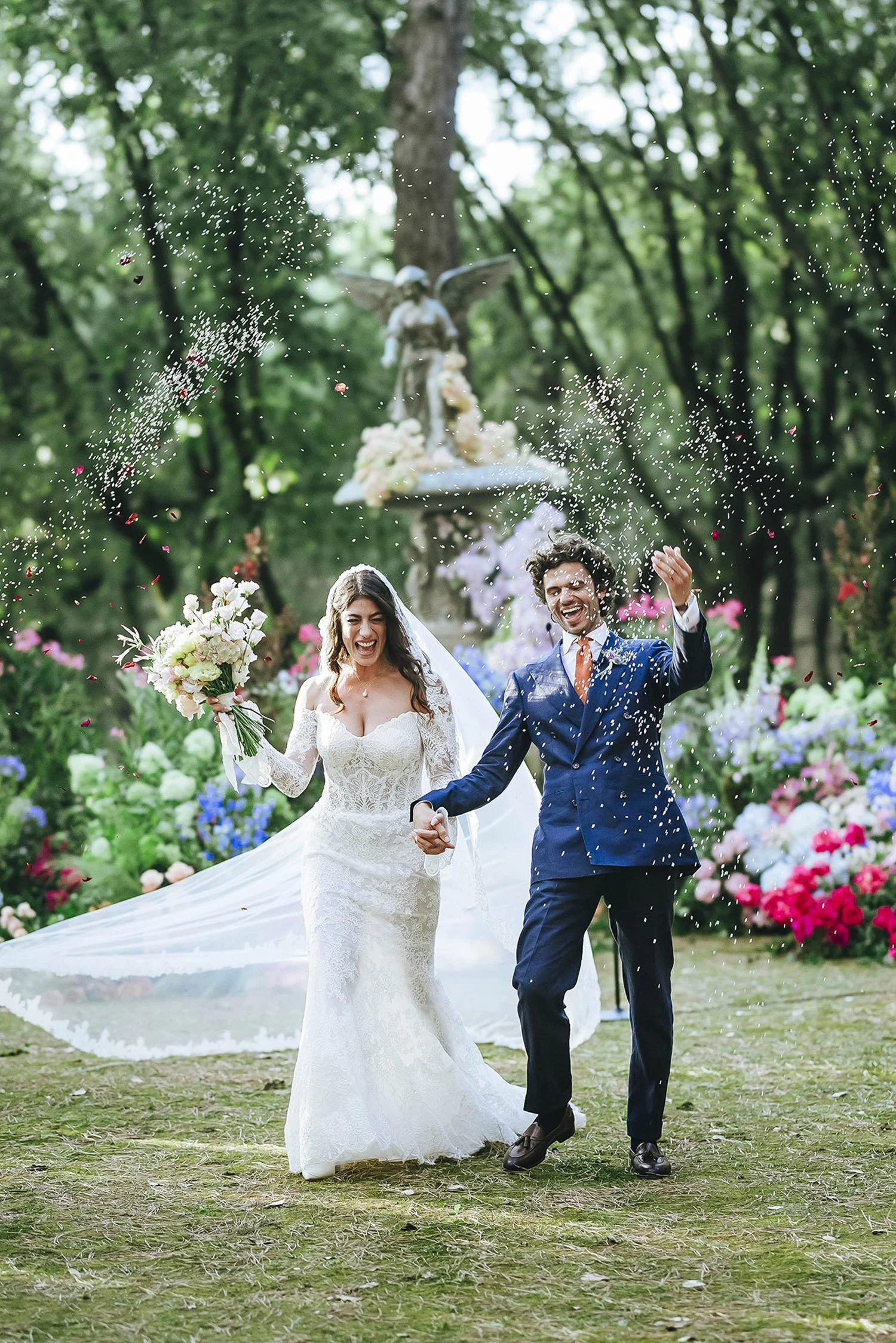 A newlywed couple celebrating outdoors, holding hands, with the bride holding a bouquet and the groom making a joyful gesture, while confetti falls around them in a garden setting with colorful flowers and green trees in the background.