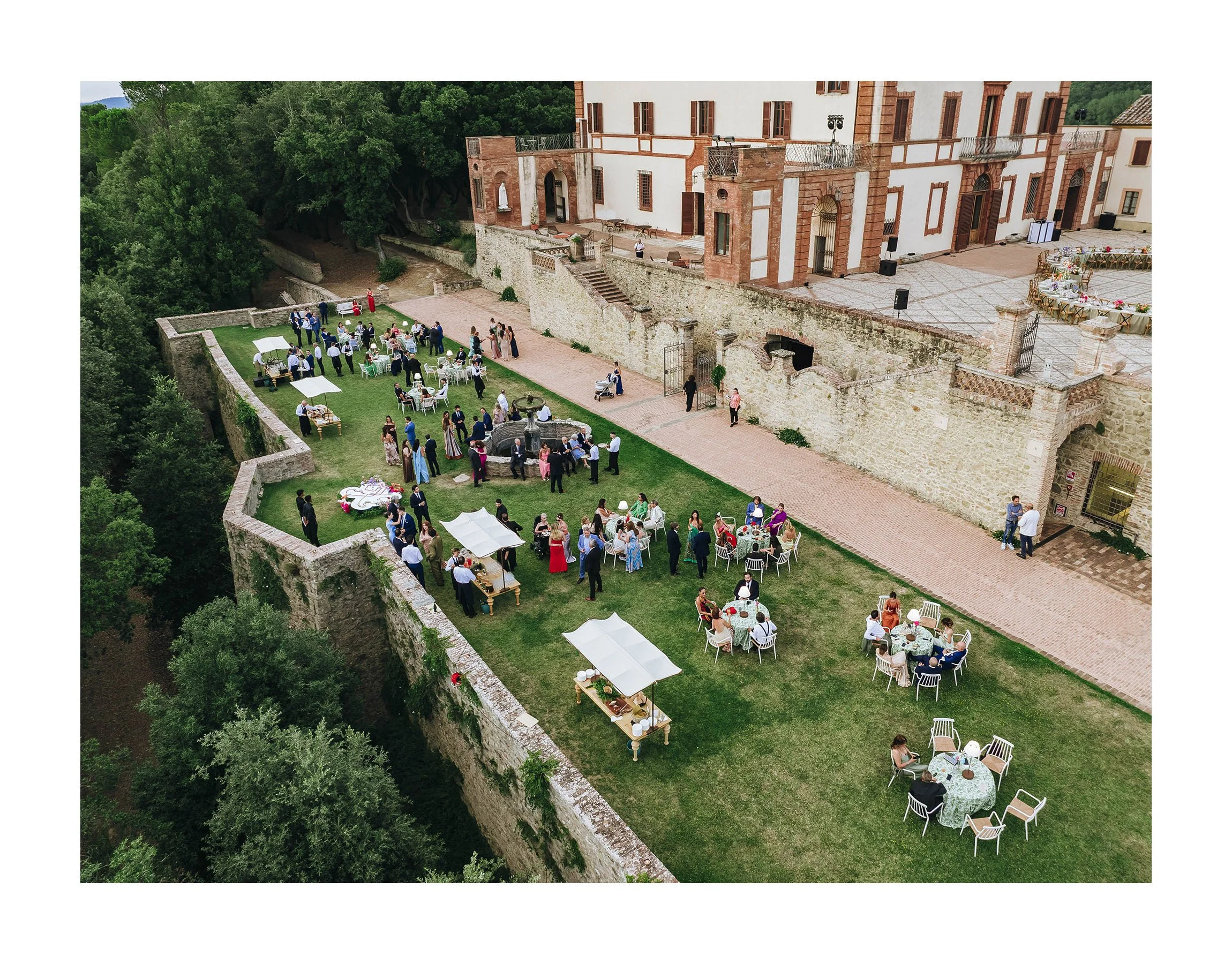 An aerial view of a wedding reception on a grassy area next to a historic stone building, with tables, chairs, and groups of people celebrating.