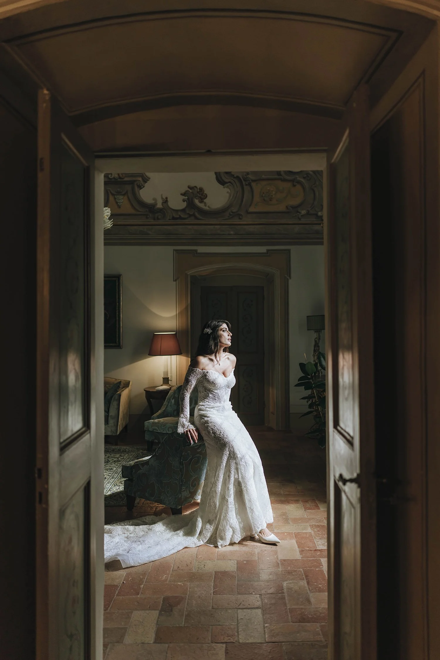 A woman in a white wedding dress sitting on a chair in a vintage styled room, framed by an open doorway.