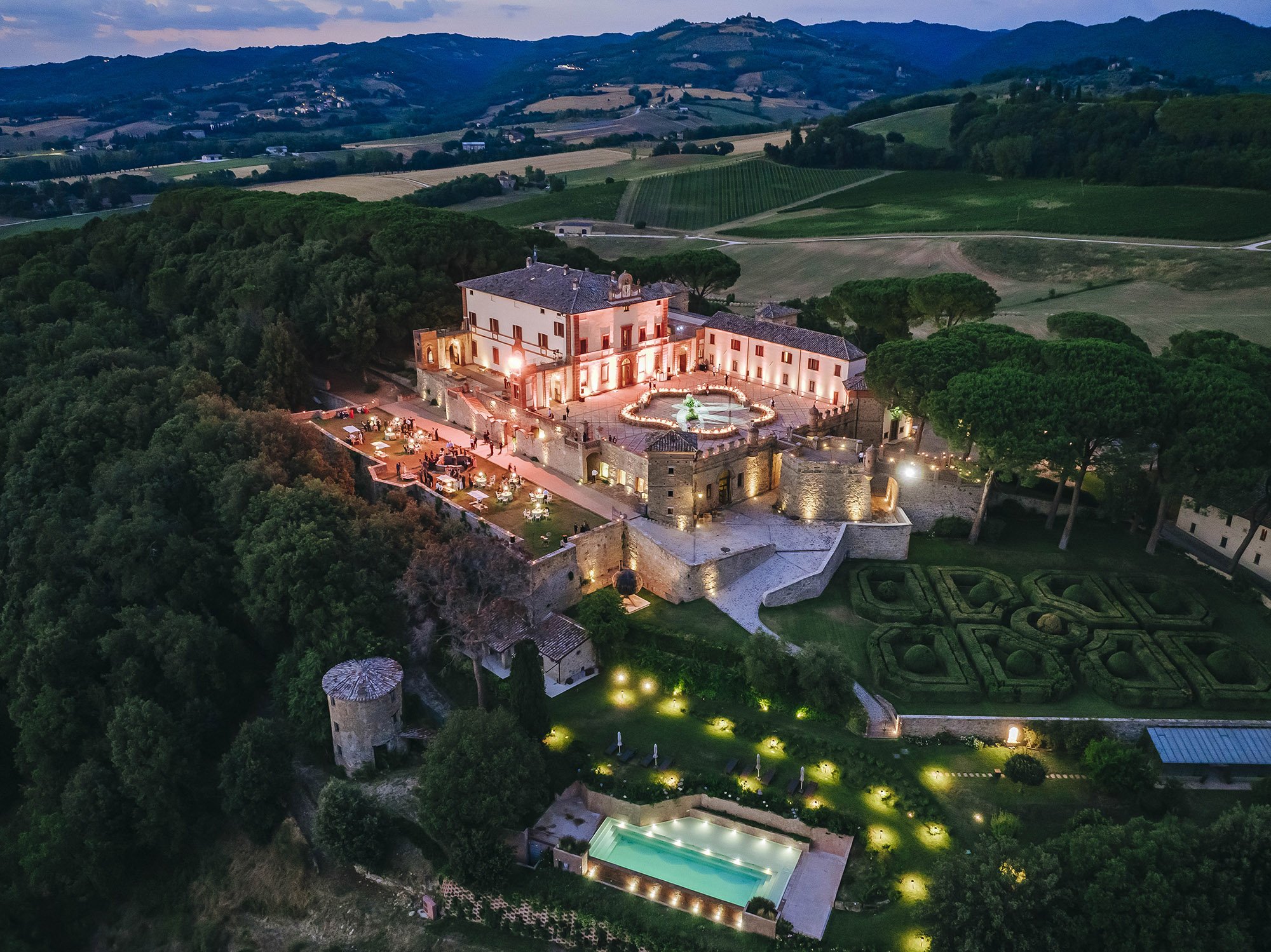 Aerial night view of a historic castle or villa with illuminated towers, surrounded by lush greenery and trees, featuring a large central square with a fountain, outdoor dining area, and a swimming pool. Rolling hills and farmland are visible in the 