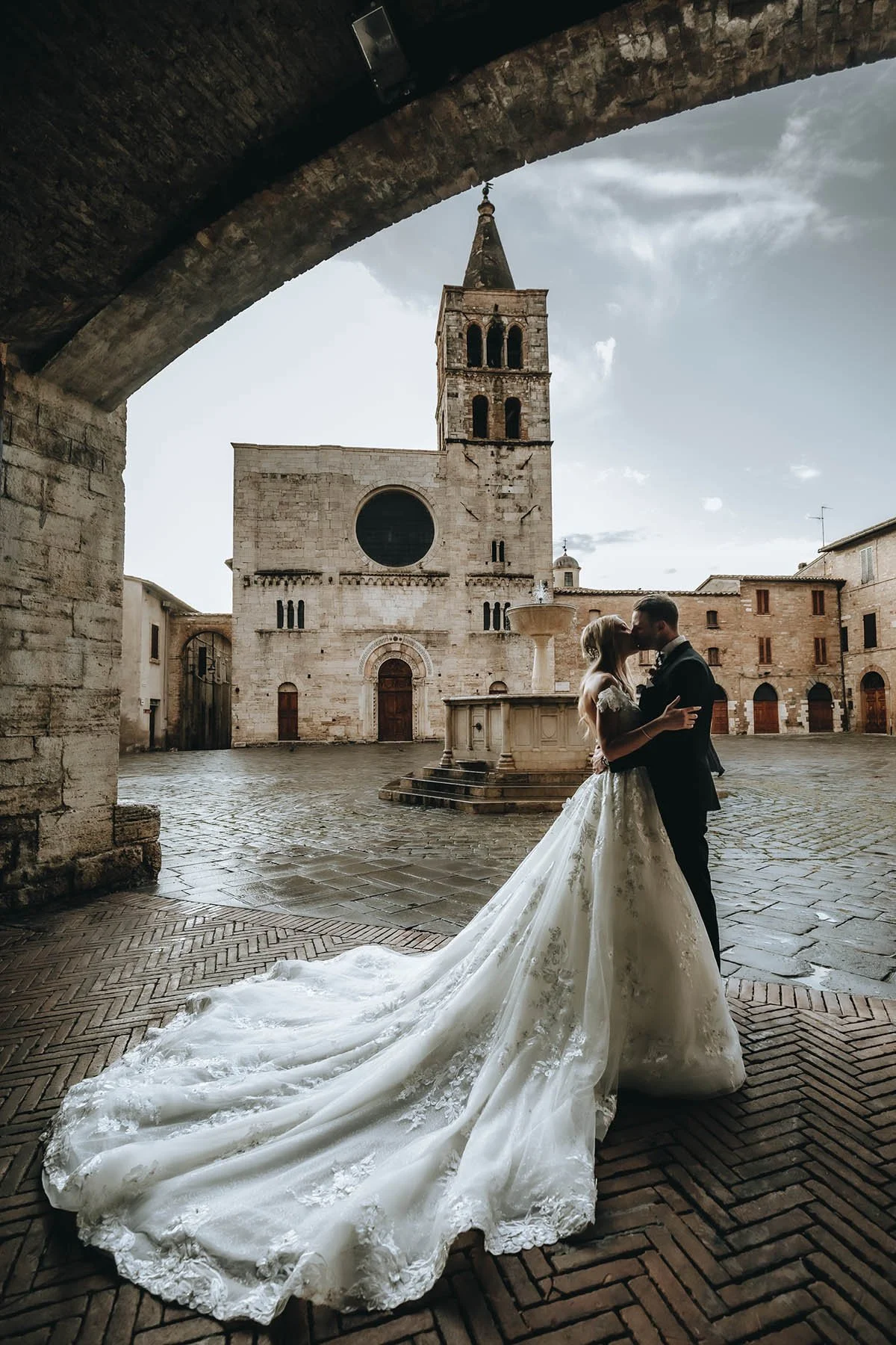 A bride and groom embracing and kissing in a historic stone town square, with an old church and tower in the background, captured during daytime with soft natural light.