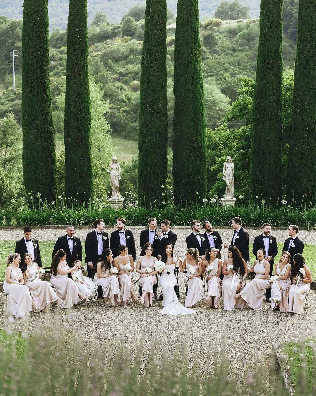 bridal party posing with bridesmaids sitting on chairs and groomsmen standing with cypresses in the background