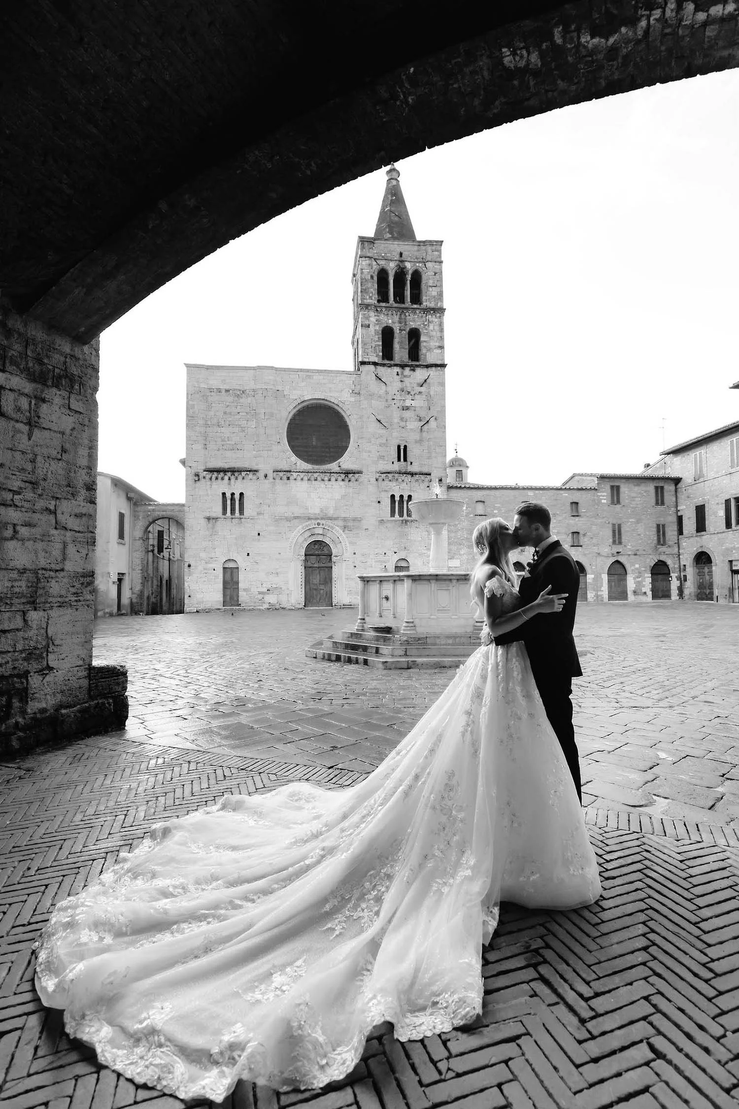 bride and groom kiss each other under an arch in an empty square in Umbria, with a medieval building in the background
