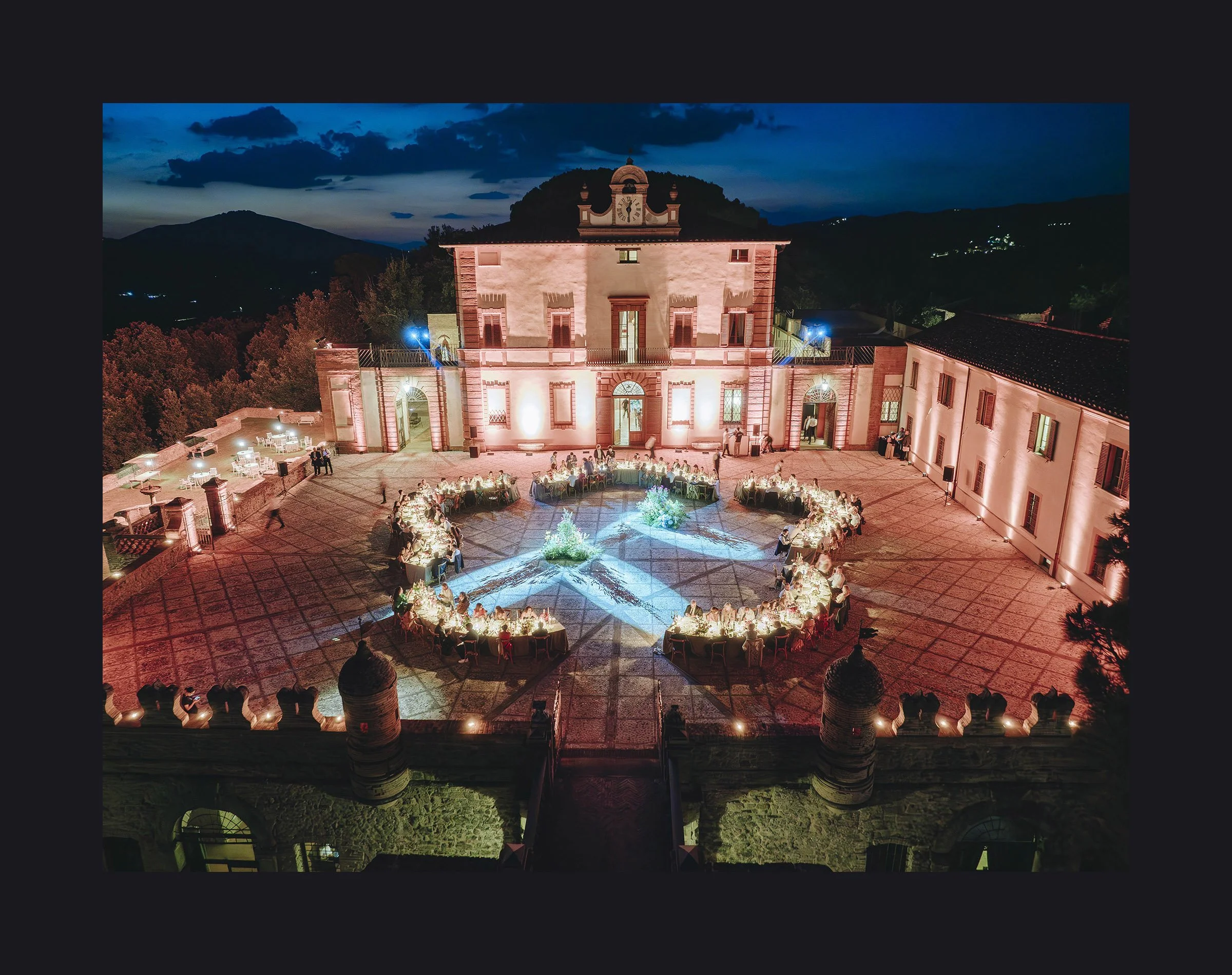 Nighttime outdoor event at a historic building with illuminated courtyard and round tables, guests seated, decorative lighting, and surrounding mountainous landscape.