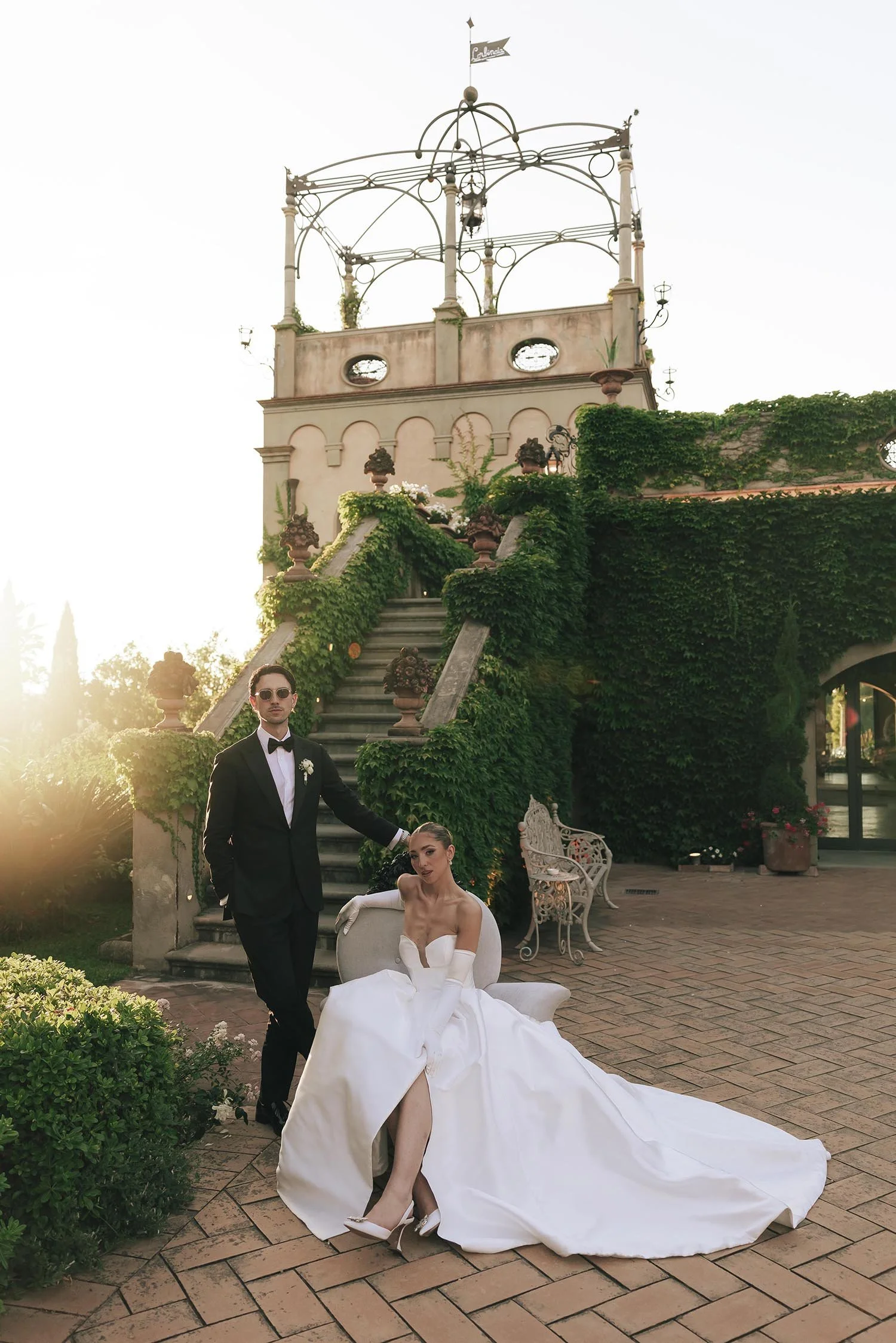 bride sitting on an armchair and groom standing beside her with the sun setting on Tenuta Corbinaia