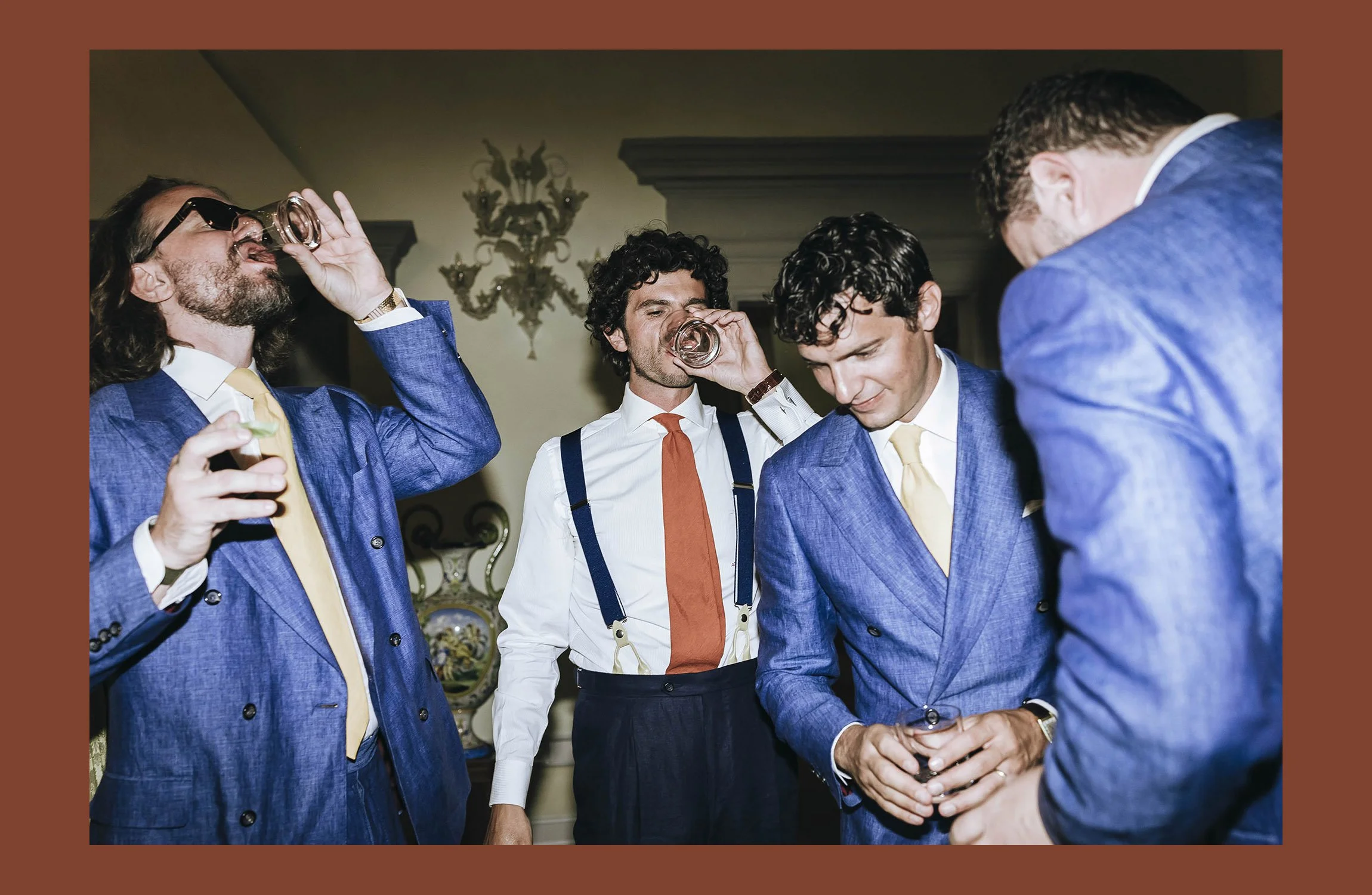 Group of four men in suits at a party, three drinking from glasses, one looking down, in a room with chandelier and decorative plates.
