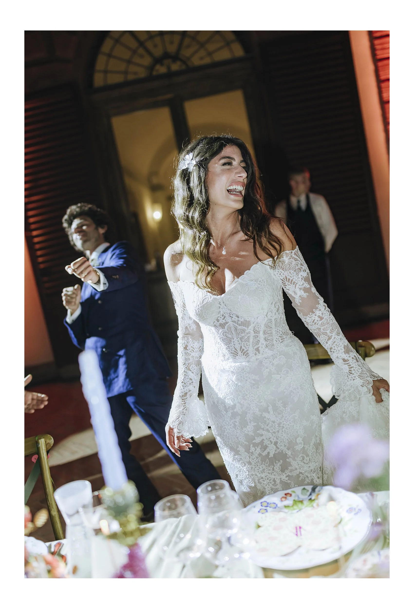A woman in a white lace wedding dress dancing and smiling at a wedding reception, with a man in a blue suit dancing in the background.