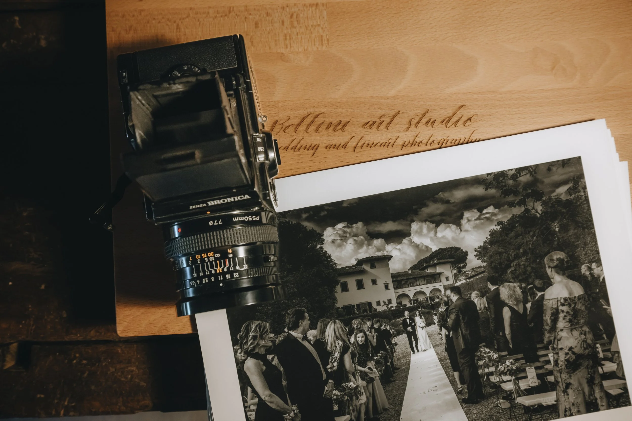 A vintage camera placed on a table next to a black and white photo of a wedding ceremony outdoors at a large house with cloudy sky in the background. Analogue film photographers in Italy