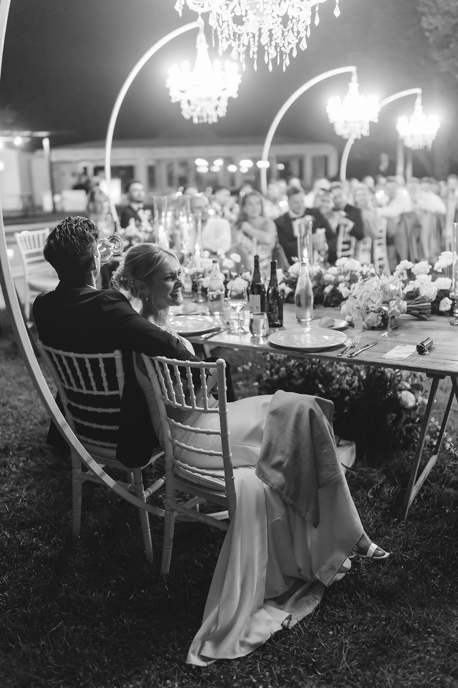 bride and groom sitting at the dinner table enjoying the party with the guests seated al fresco under chandeliers