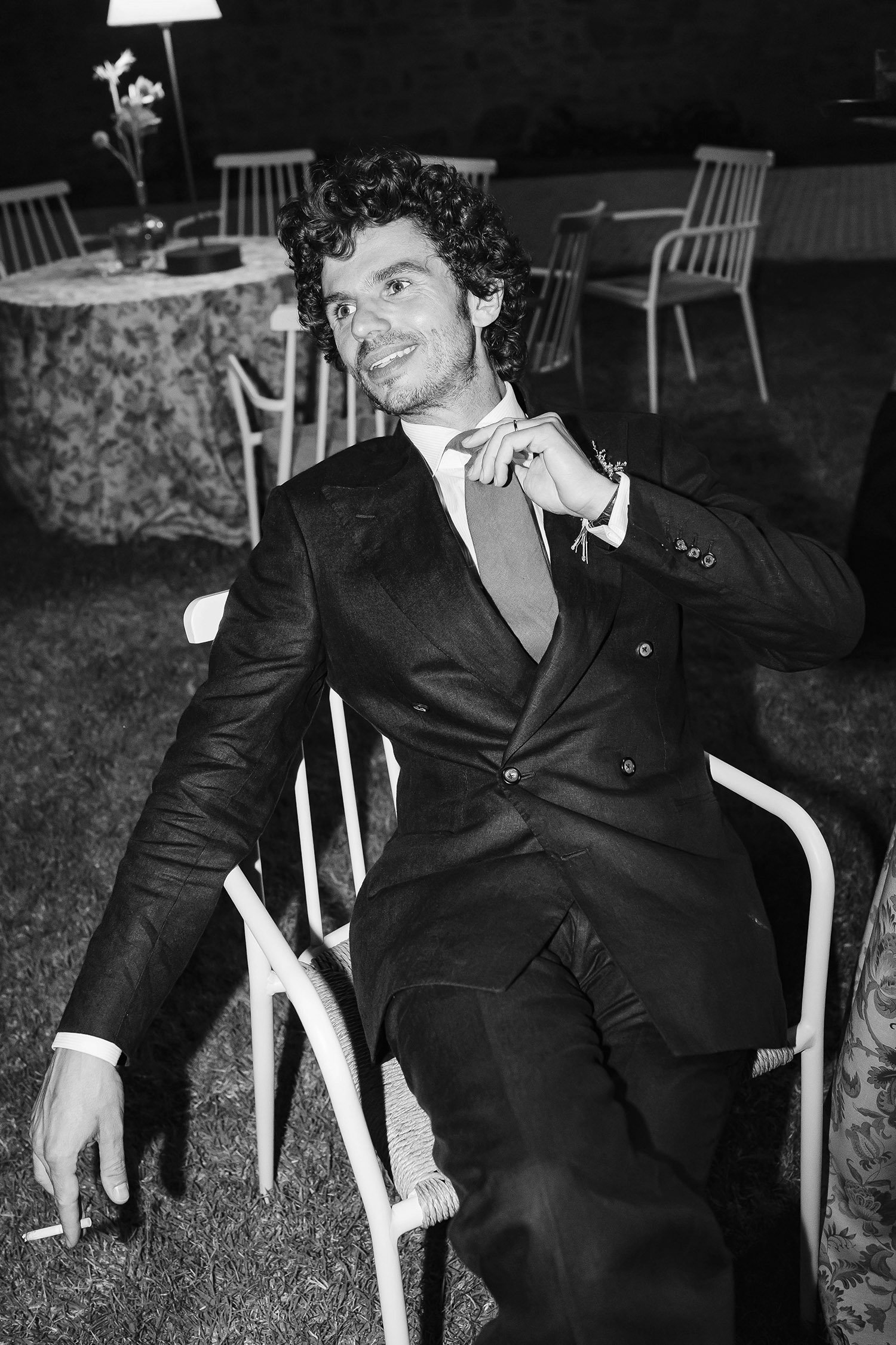 A man with curly hair sitting on a white chair at a table, wearing a dark suit and holding a cigarette, smiling and looking to his right at a nighttime outdoor event.