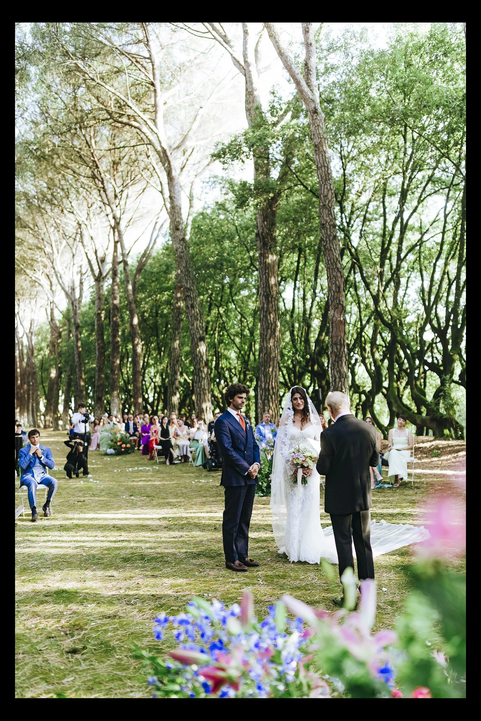 A wedding ceremony taking place outdoors in a forested area with tall trees and green foliage. The bride and groom are standing in front of an officiant, with guests seated behind them. The bride is wearing a white wedding dress and holding a bouquet