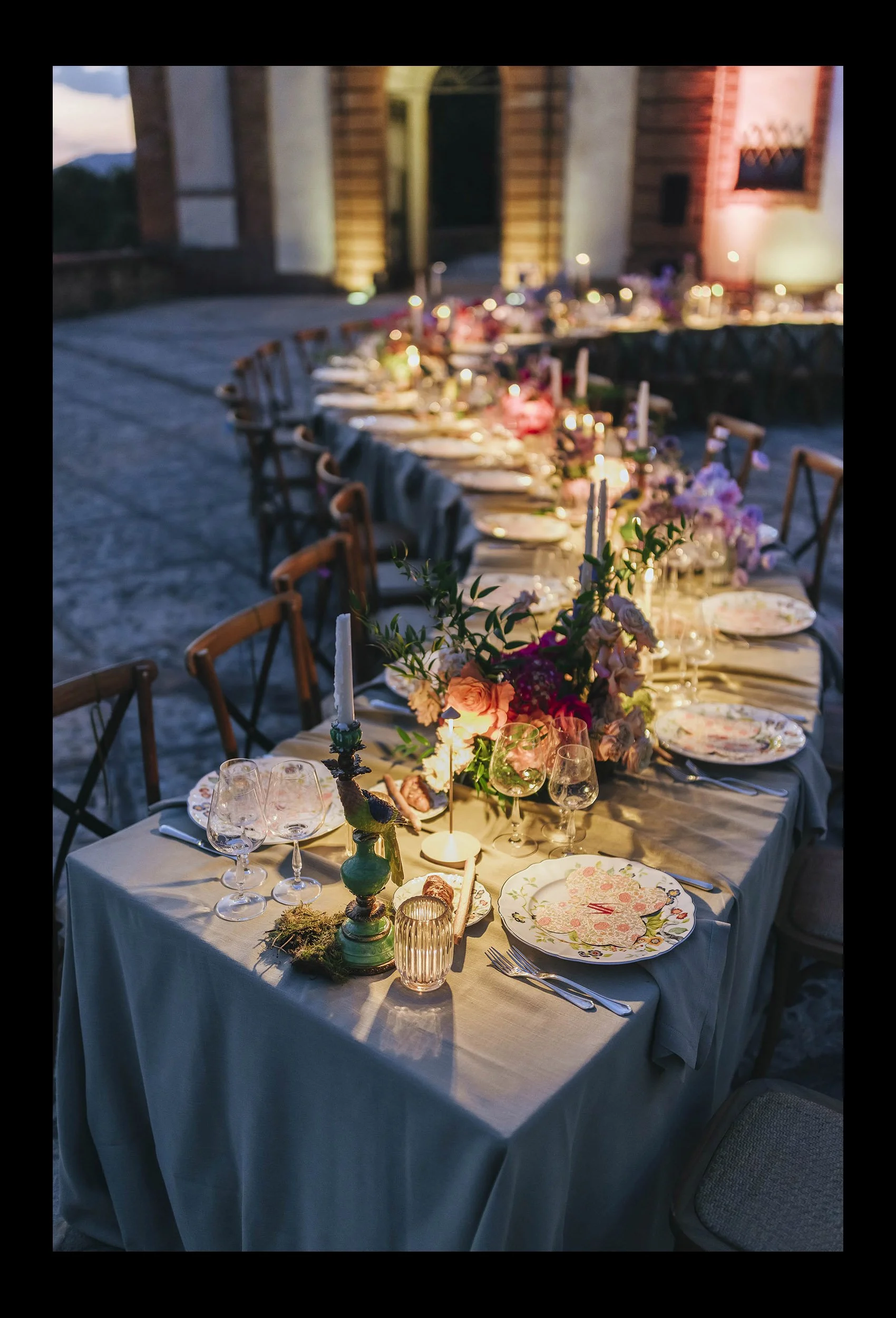 Evening outdoor banquet table decorated with floral arrangements, candles, and fine china, set for a formal event.