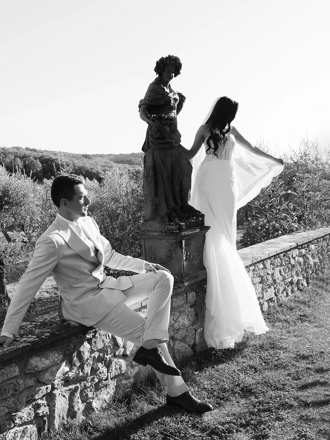 The bride standing and holding the veil while the groom is sitting under an ancient statue