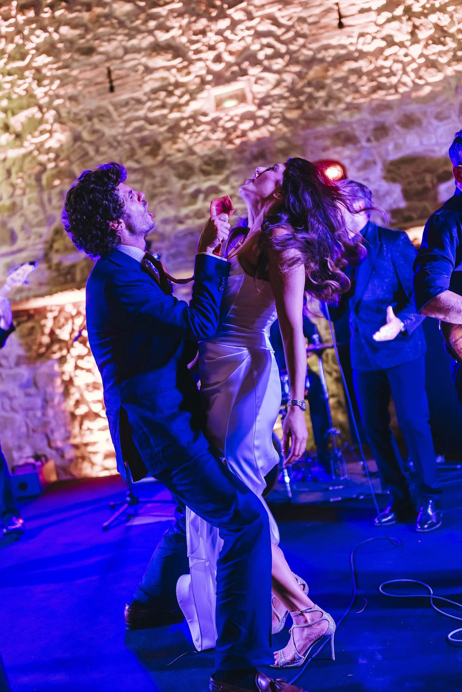 A man in a suit and a woman in a white dress perform a dance or romantic moment on stage against a stone wall, with other people watching or participating nearby, and stage lighting creating a lively atmosphere.