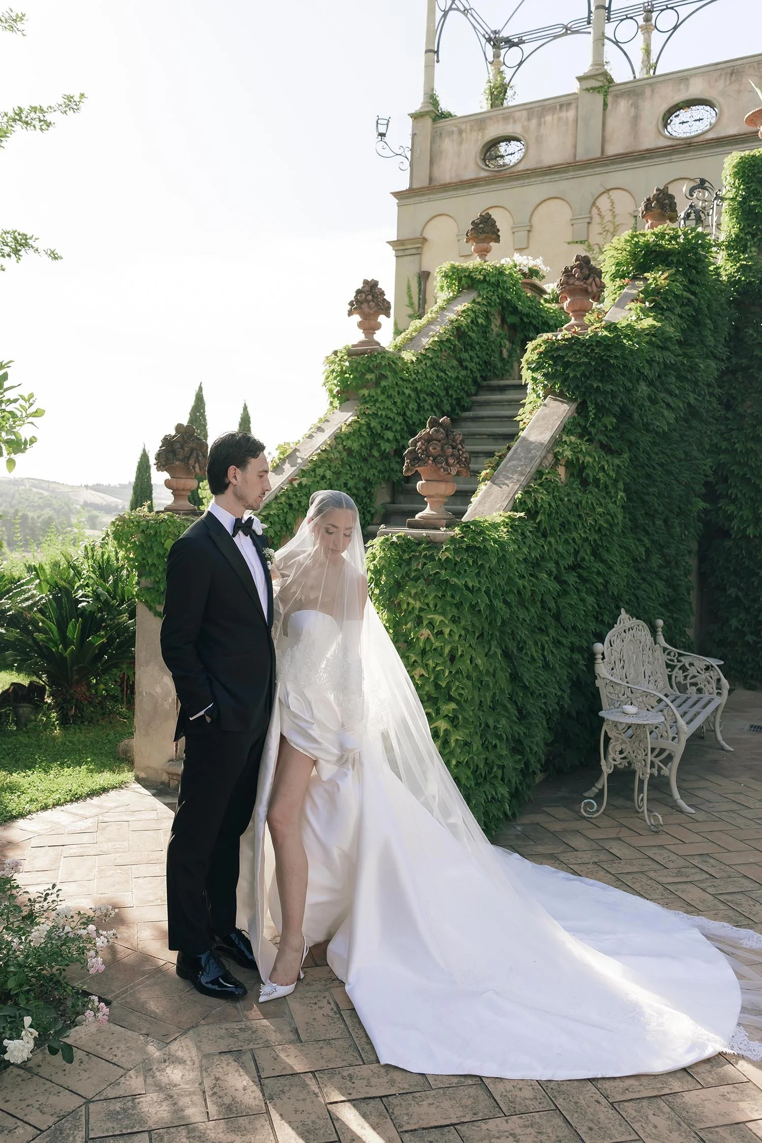 bride under the veil fixing her skirt with the groom standing beside her and the sun setting behind Tenuta Corbinaia