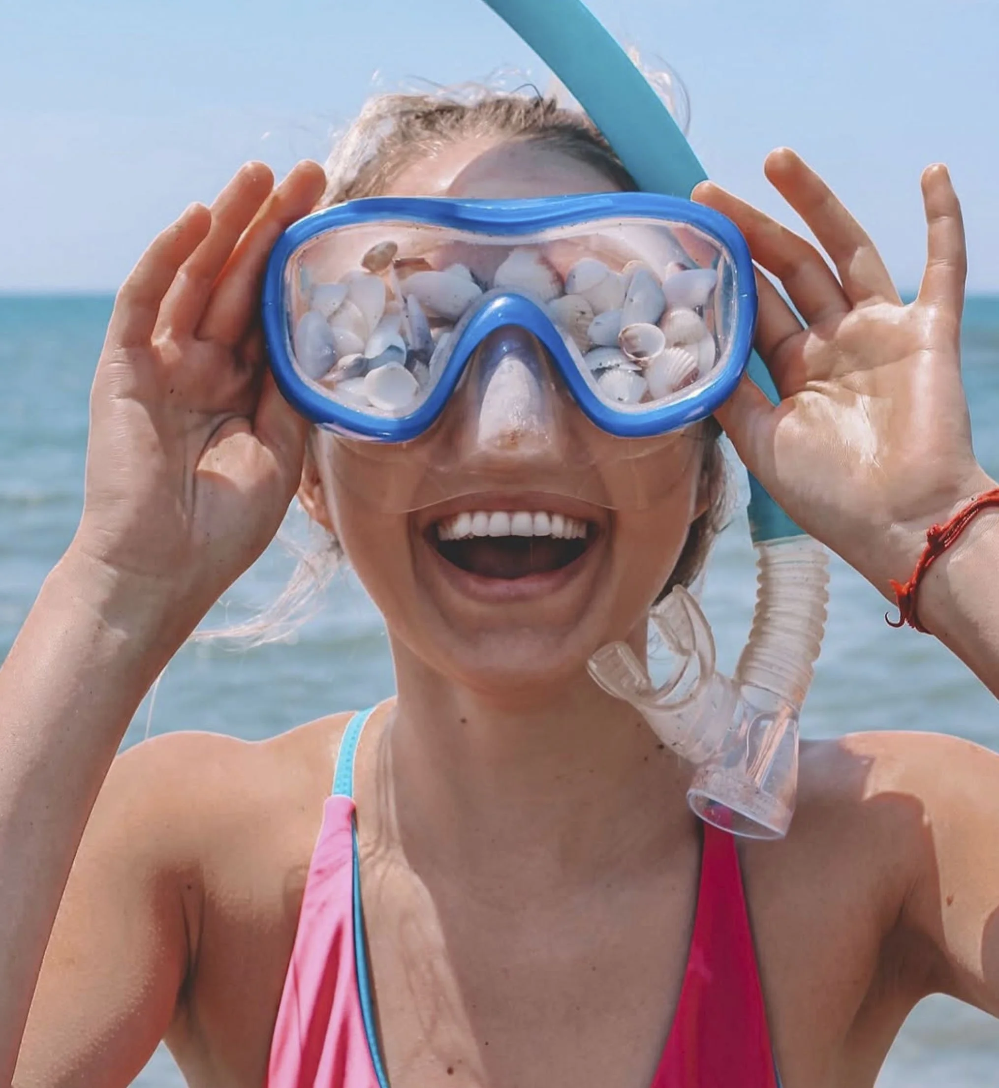 A woman at the beach wearing swimming goggles filled with seashells, smiling with open mouth.