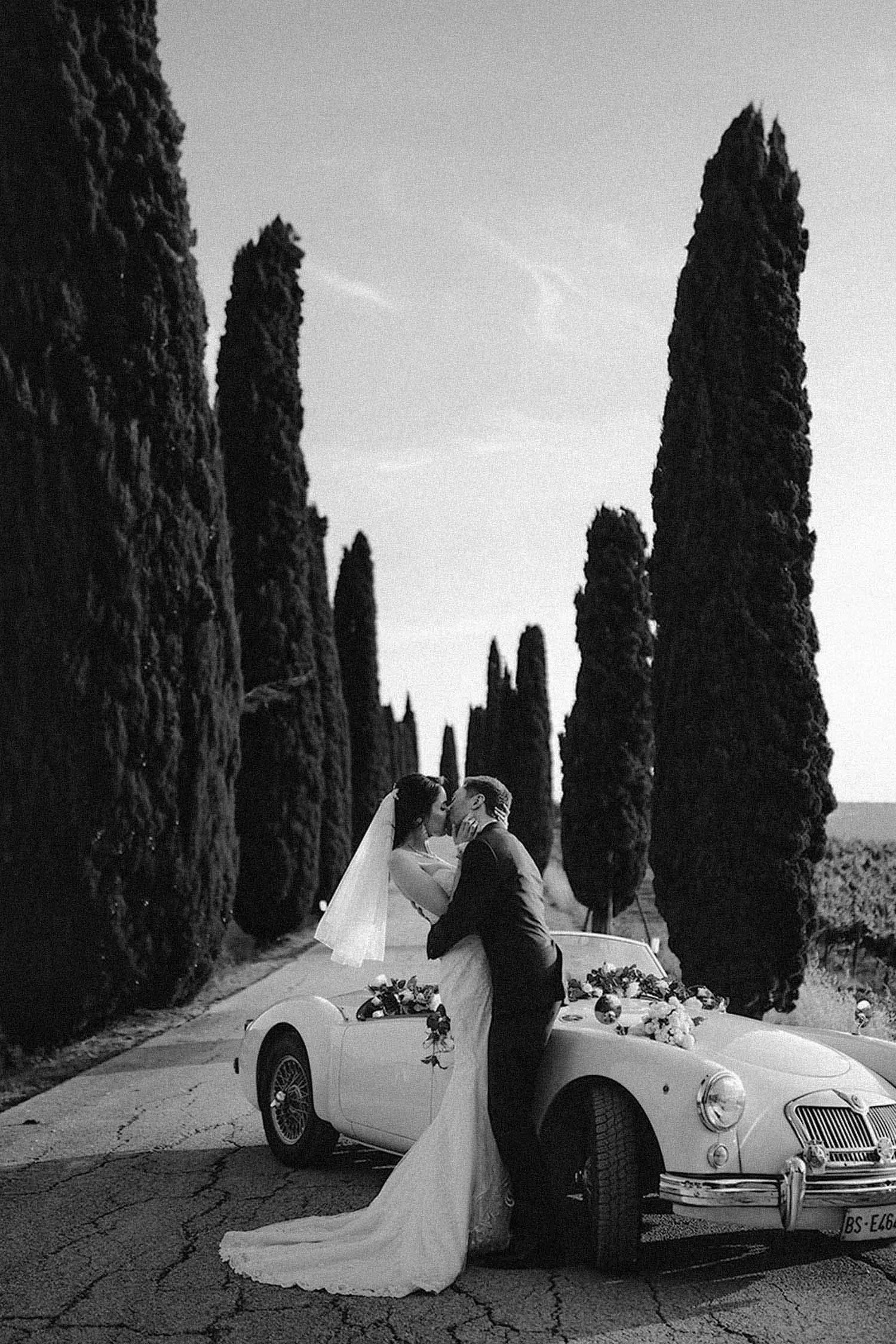 Bride and groom kissing outside of a vintage italian car along a cypresses road in a rural italian countryside.  photo by Rellini art studio best wedding photographers in Italy