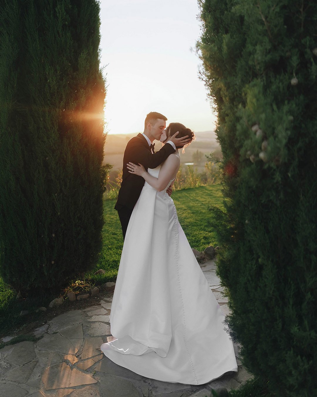 Bride and groom kissing each other with the sun setting behind the cypresses