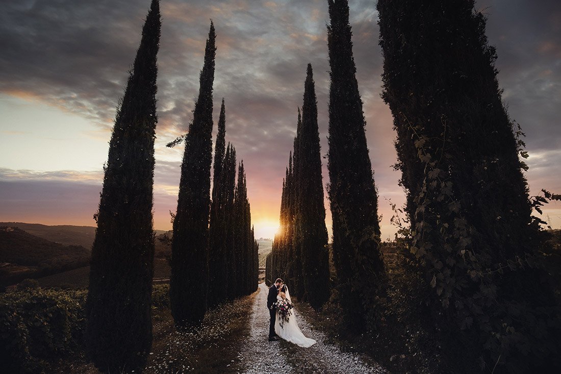 A couple in wedding attire hugging on a path between tall cypress trees at sunset.