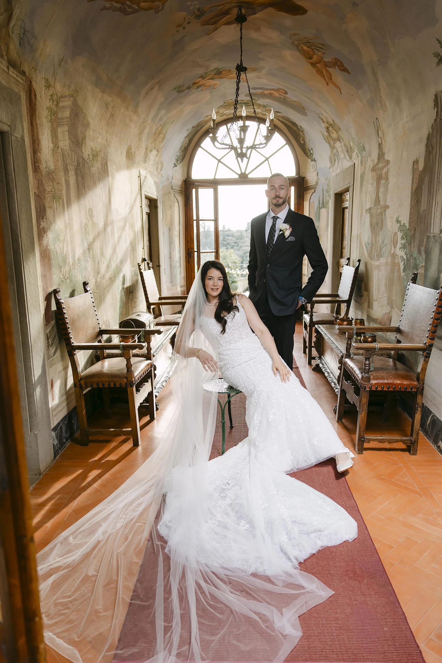 Timeless portrait of bride sitting on a chair and groom standing beside her in the gallery with frescoes at Villa Medicea di Lilliano in Florence