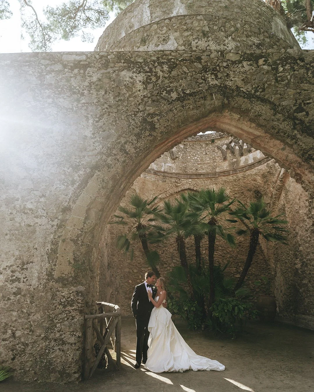 bride and groom holding each other under a roman building at Villa Rufolo in Ravello