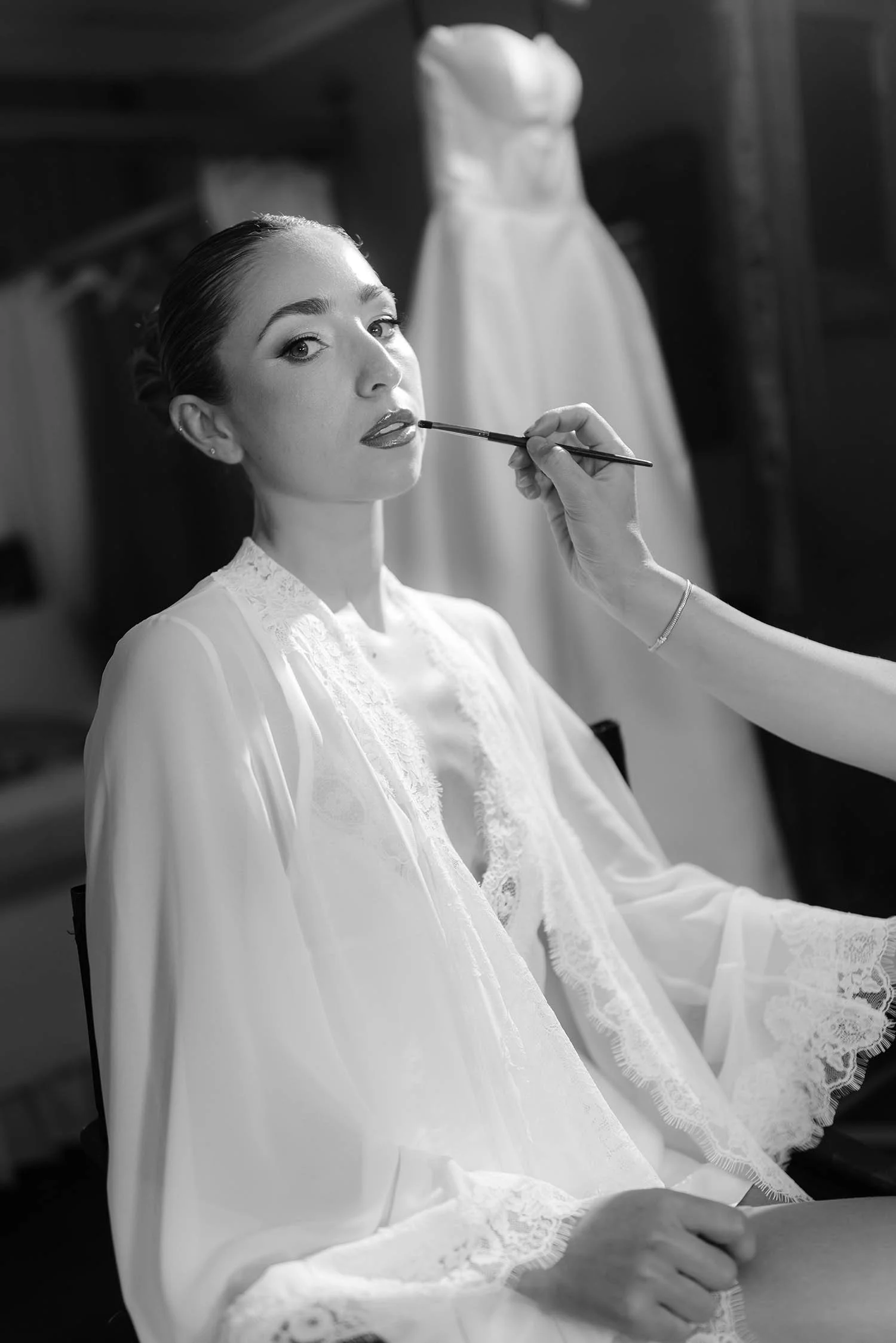 bride receiving her makeup while sitting in the bridal suite with the dress hanging in the background