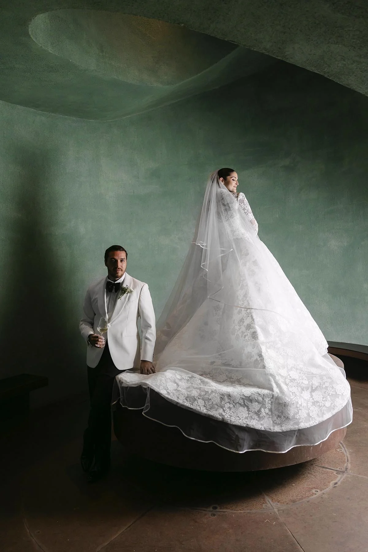 Elegant editorial wedding portrait of Ricardo and Norimar, featuring a bride in a dramatic lace gown and veil alongside the groom in a white tuxedo, surrounded by sculptural architecture and soft natural ligh