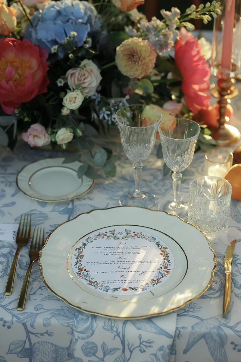 A beautifully set elegant table with a floral centerpiece, gold-rimmed china plates, crystal glasses, and gold cutlery. The tablecloth has a blue and white botanical pattern, and there are pink and white flowers in the centerpiece.