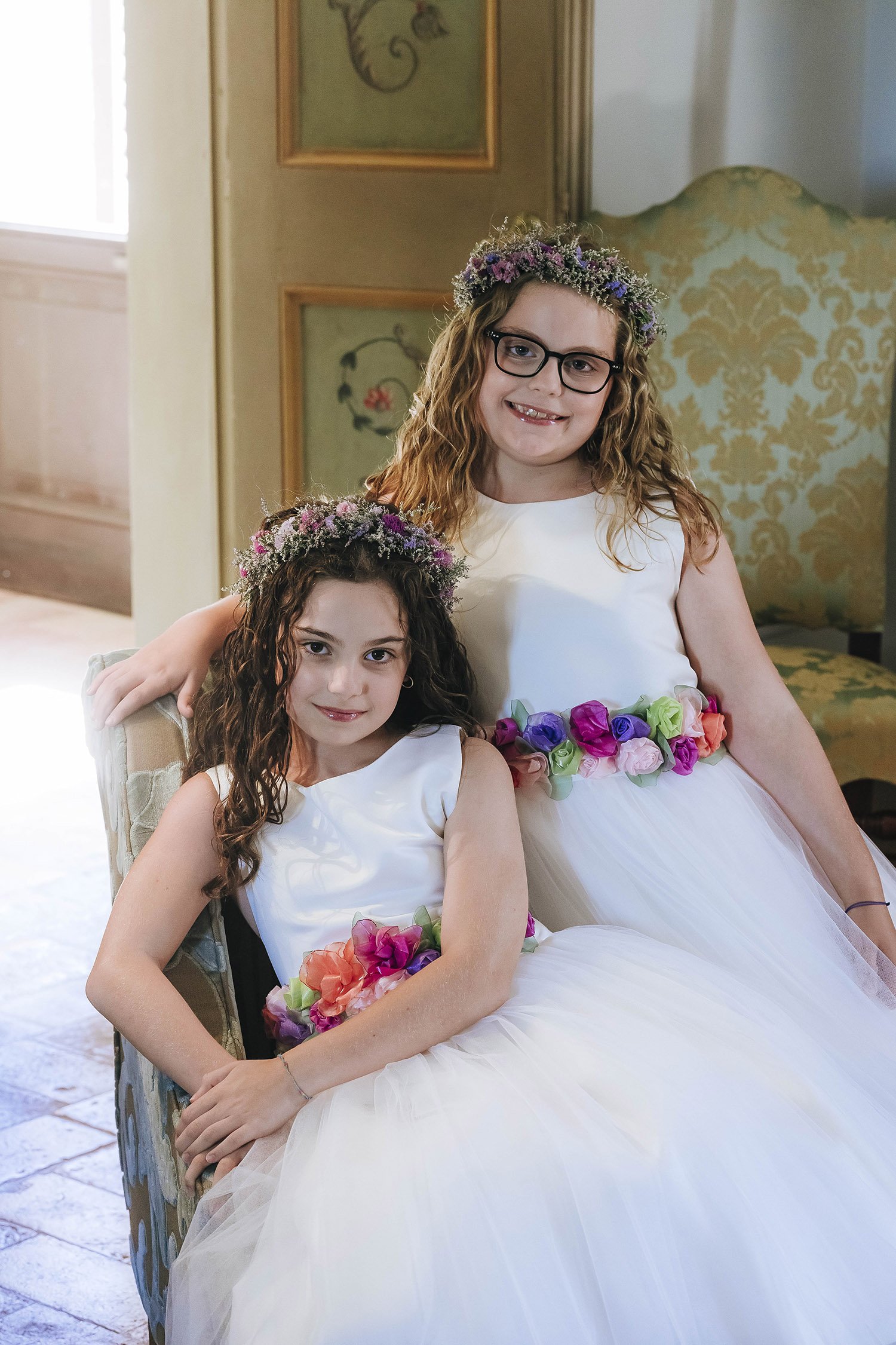 Two young girls in white dresses with flower belts and floral crowns, sitting together indoors, smiling at the camera.