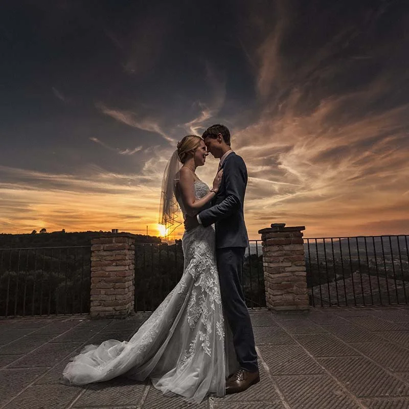A bride and groom standing close together on a balcony at sunset, embracing and touching foreheads, with a scenic view and a sunset sky in the background.