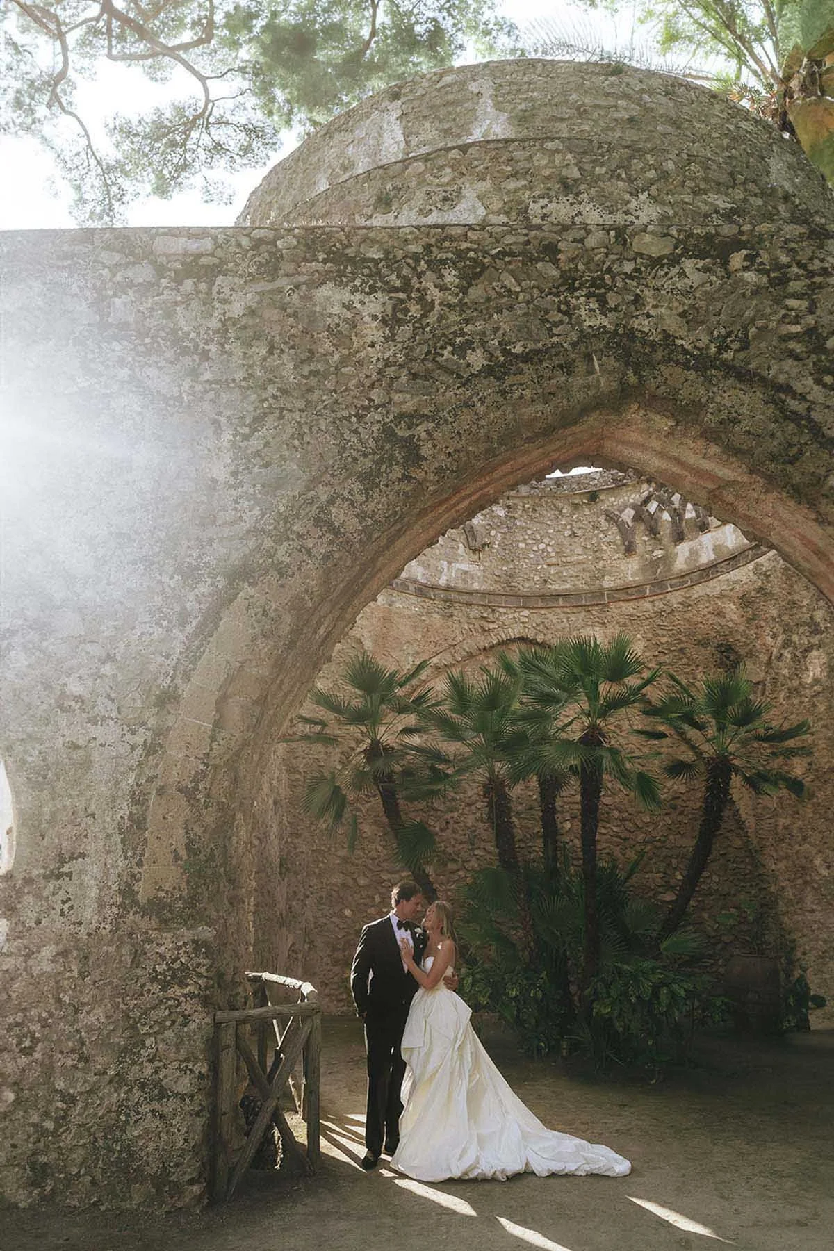 timeless portrait of bride and groom holding each other under a roman building at Villa Rufolo in Ravello with the sun setting in the background