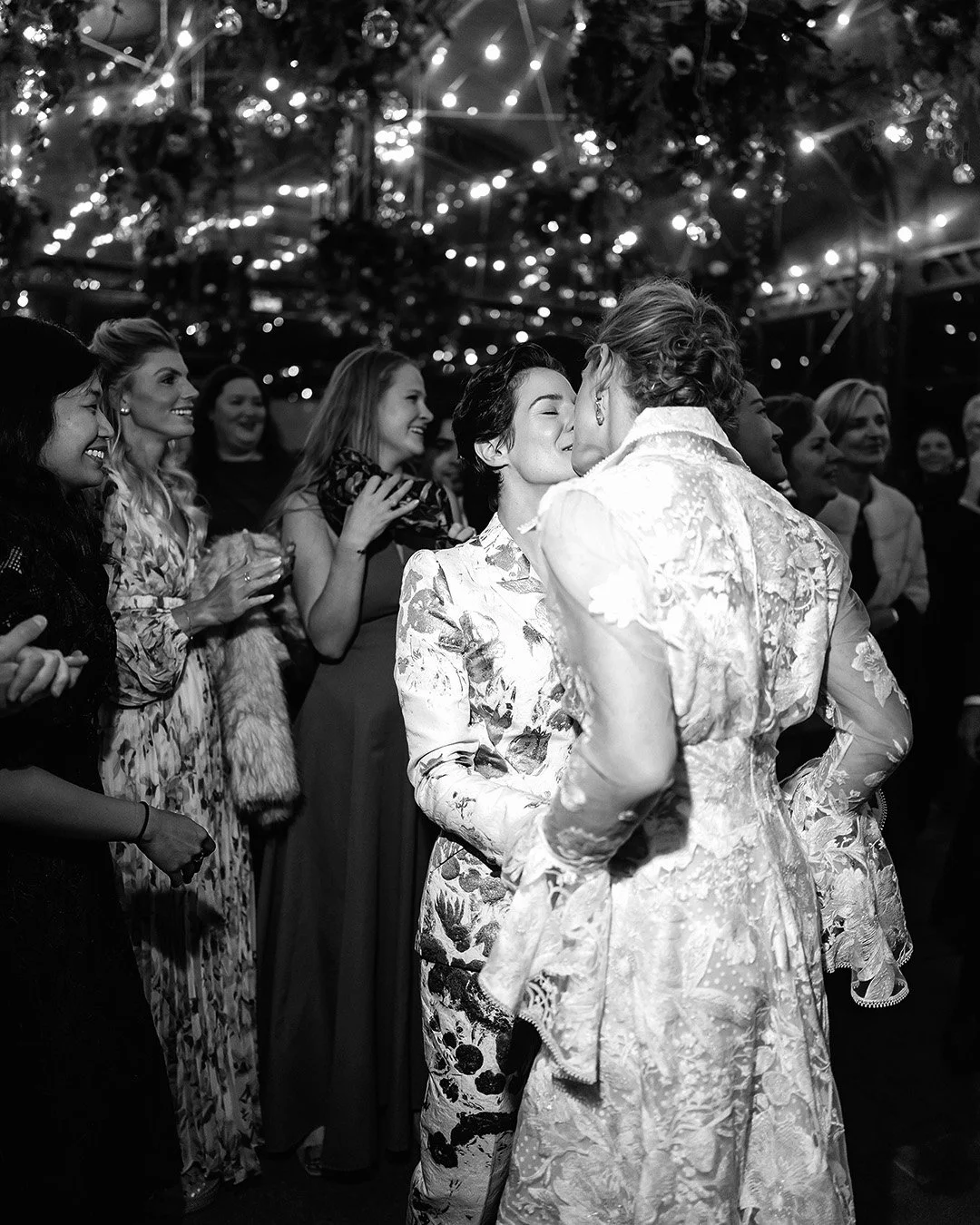 Two brides kissing with guests around during the wedding party under a marquee
