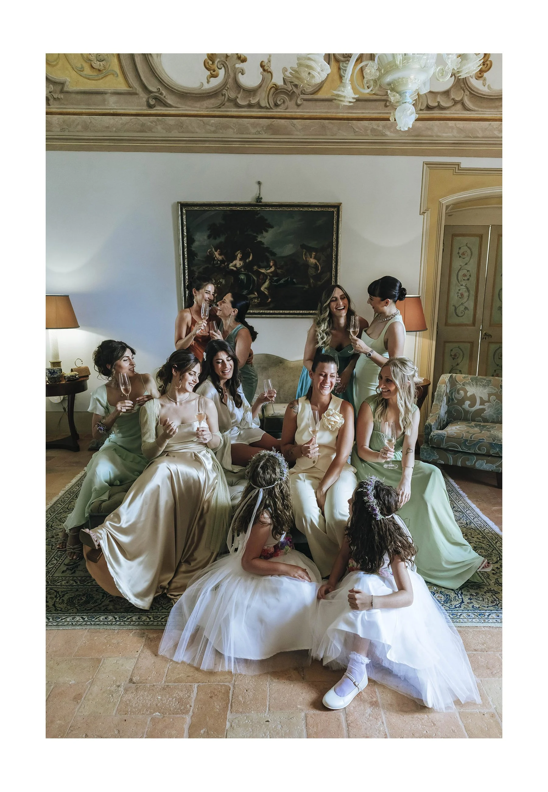 A group of women celebrating in elegant dresses, holding champagne glasses, in a luxurious room with classical decor, including a chandelier, painting, and sofas, appearing joyful and engaged in conversation.
