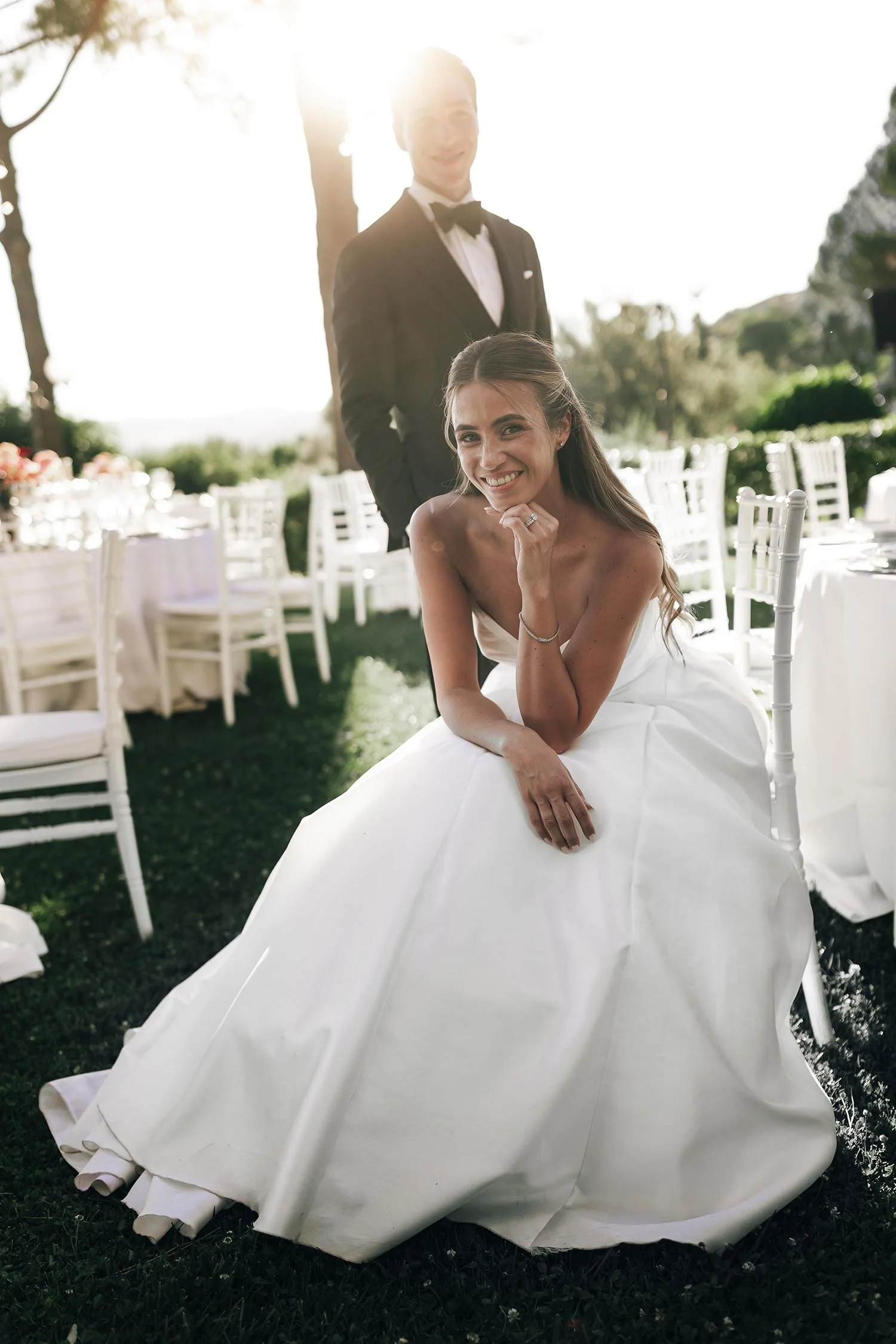 bride sitting on a chair with the groom standing in the background with the tables ready for the dinner and the sun setting behind the trees