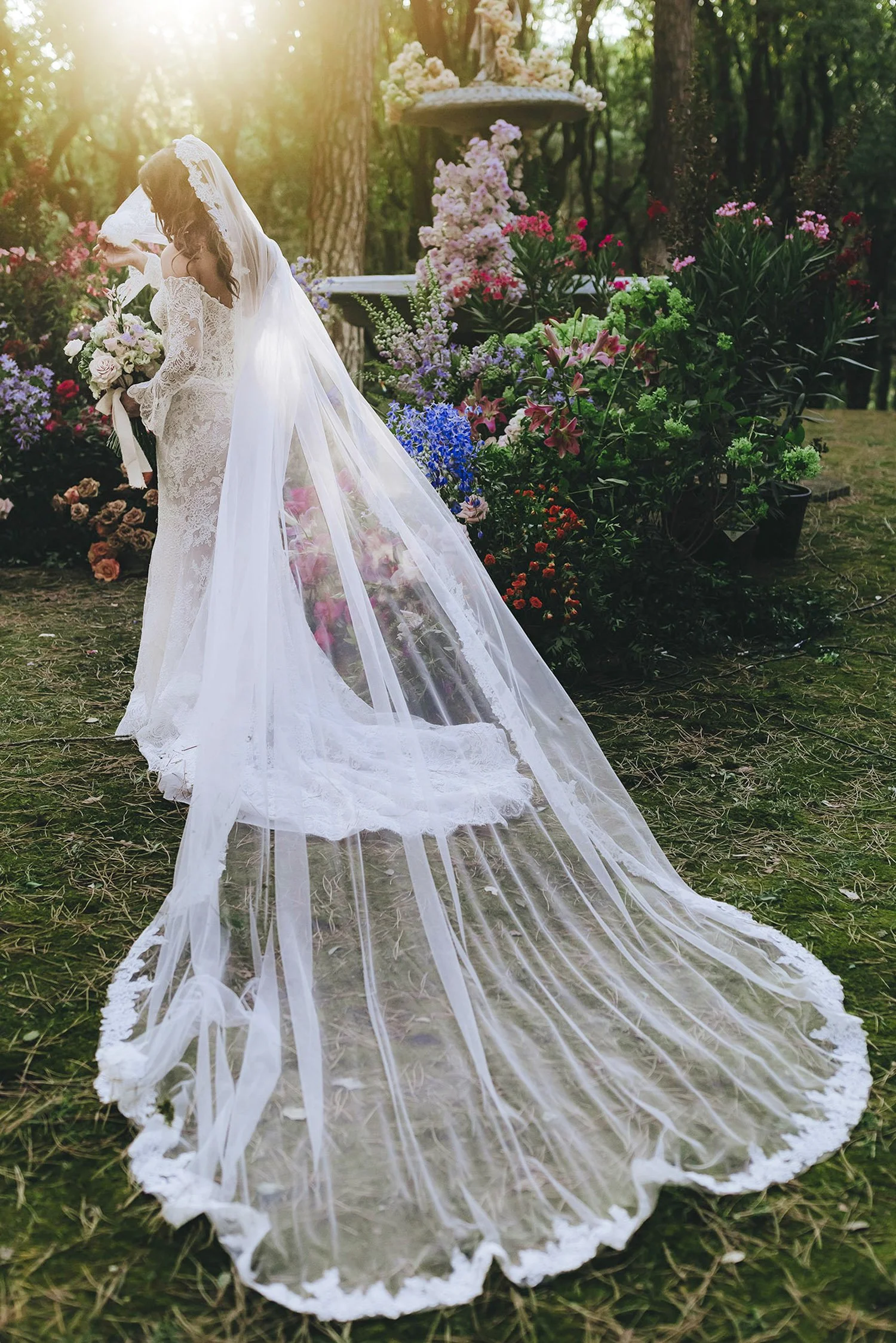 A bride in a lace wedding gown and long veil stands outdoors in a lush, flower-filled setting during sunset, holding a bouquet of flowers.