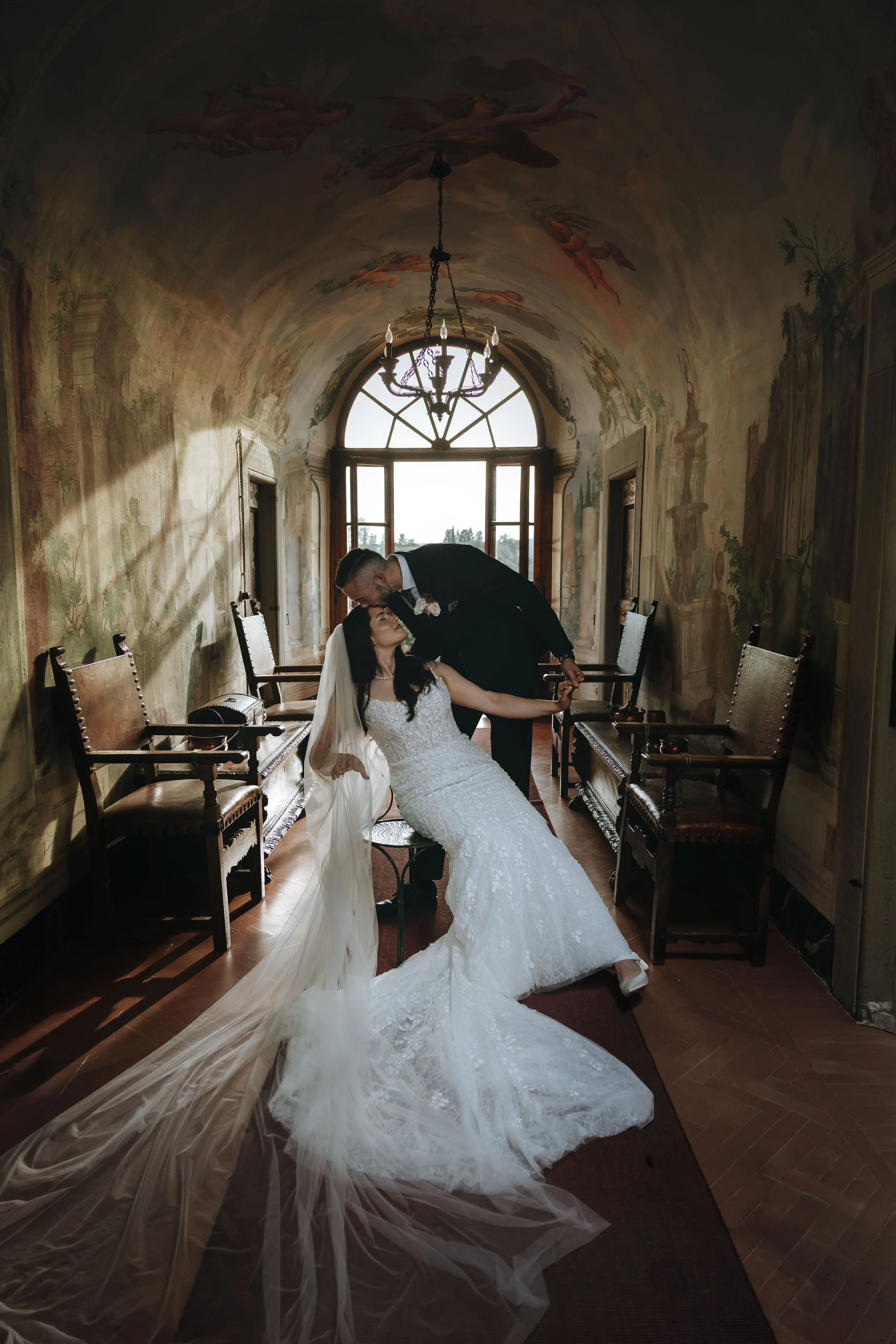 Portrait of the bride sitting on a chair in a gallery with frescoes and the groom kissing her