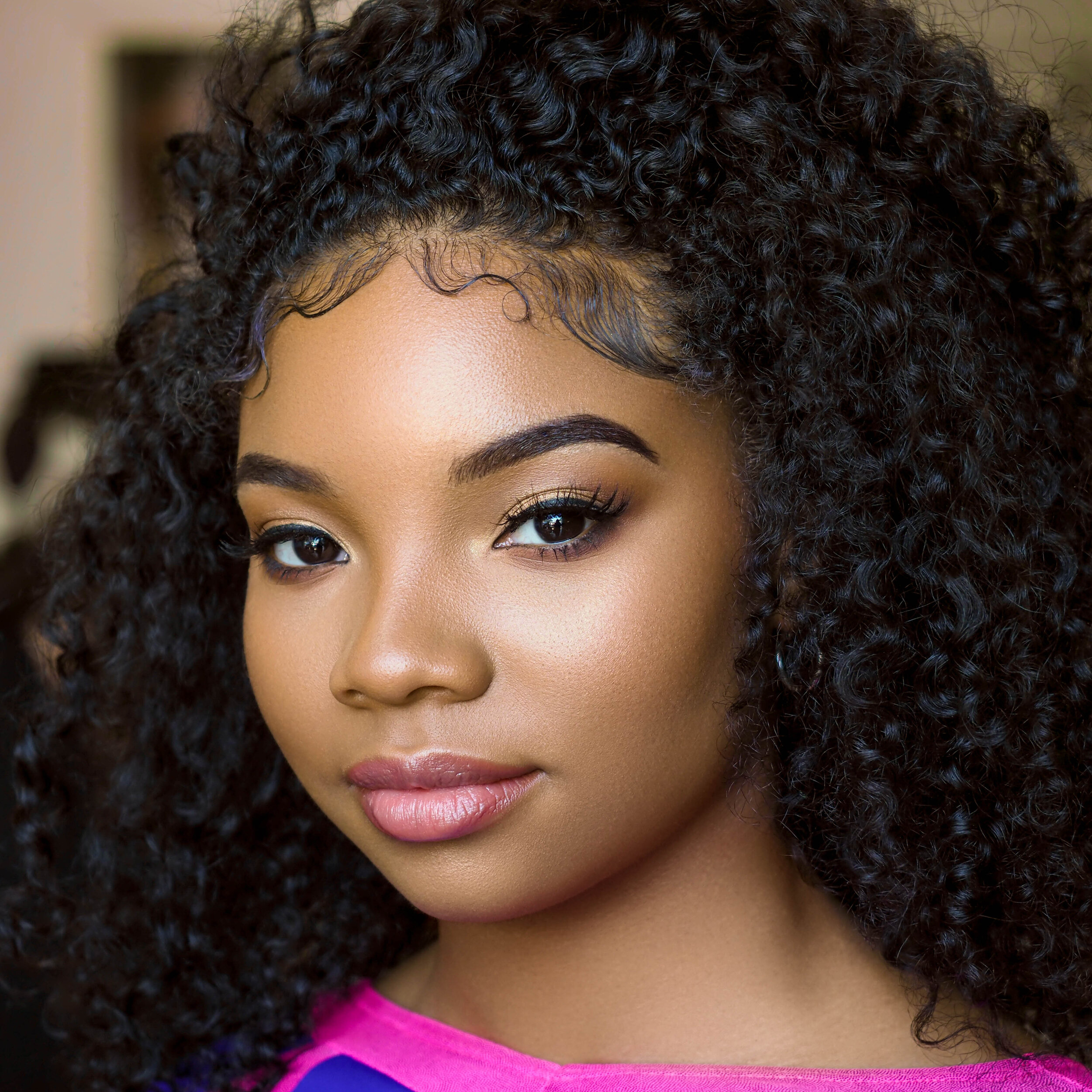 Close-up portrait of a young woman with curly black hair and makeup, wearing a pink top.