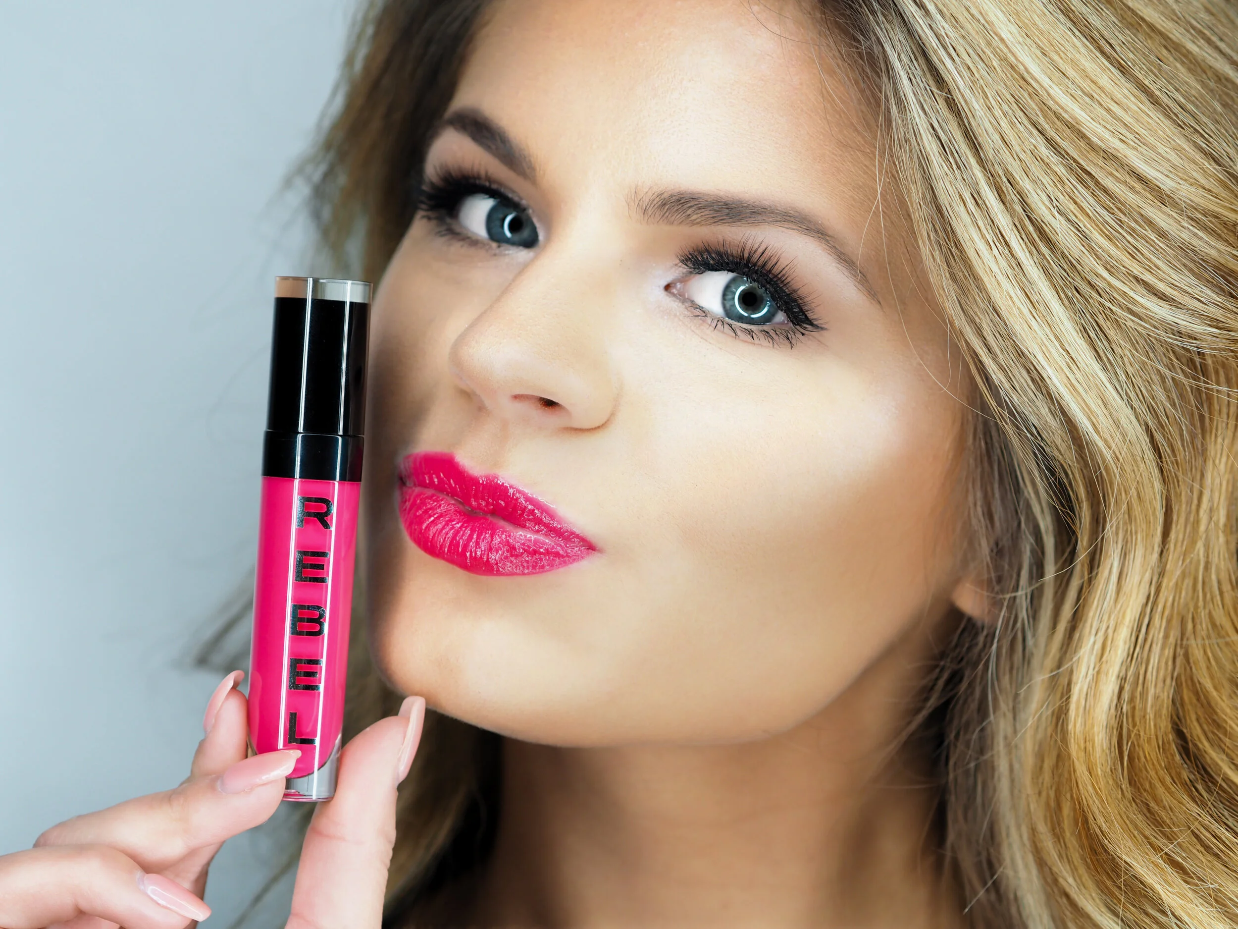 Close-up of a woman with blonde hair, blue eyes, wearing pink lipstick, holding pink lipstick tube near her face.