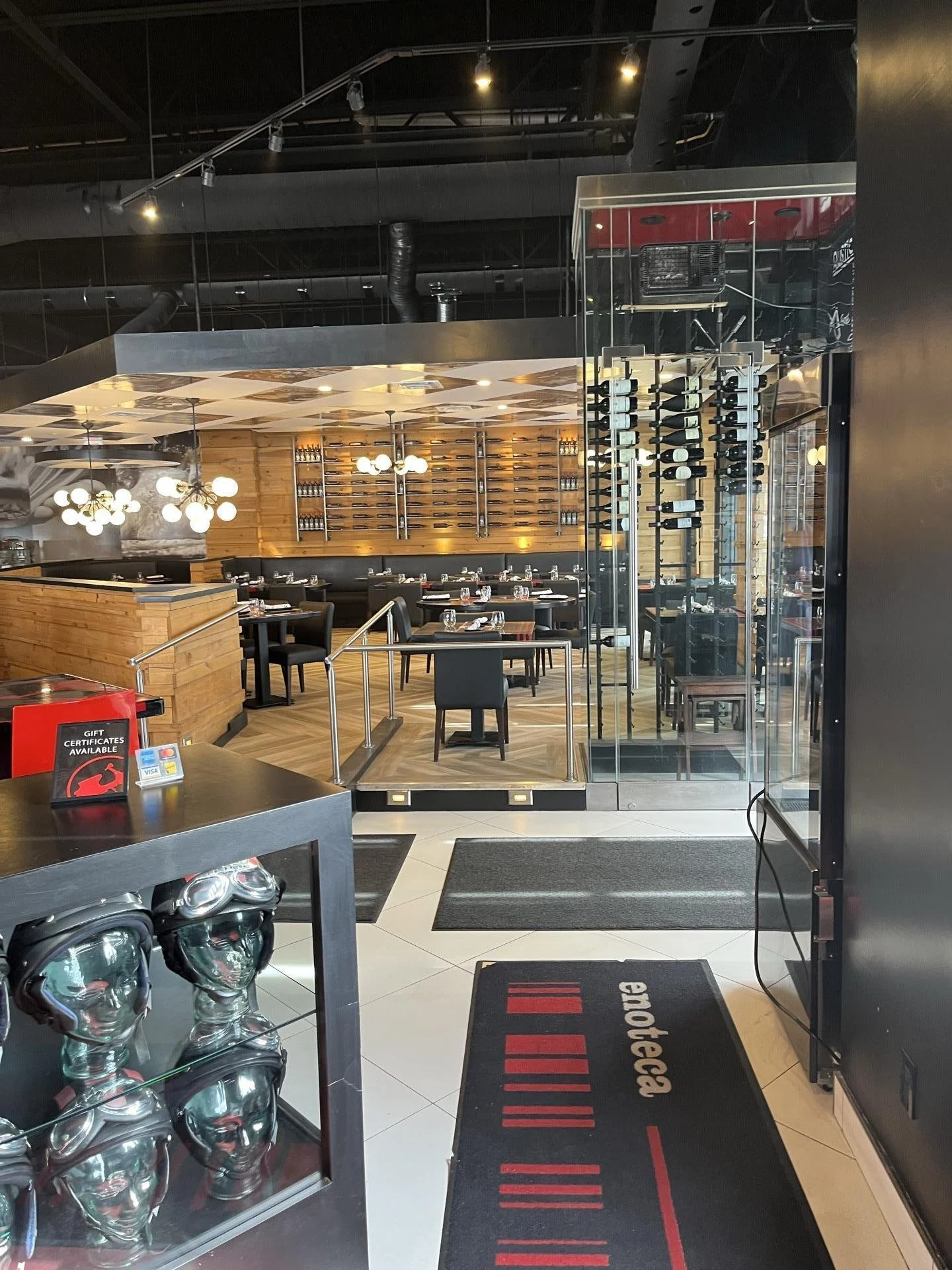 Interior view of a modern restaurant with wooden and black decor, tables set for dining, wine racks on the wall, and a glass wine storage case visible on the right.