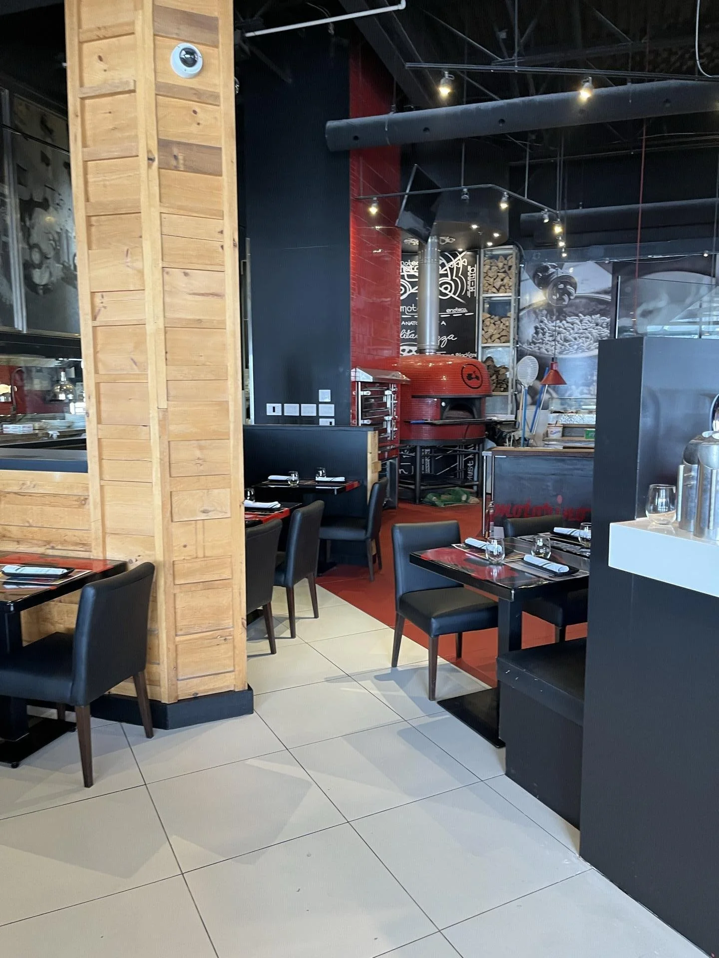 Inside a modern restaurant with black and wood decor, including black chairs, tables set with napkins and glassware, a large red pizza oven, and a wall with coffee bean artwork.