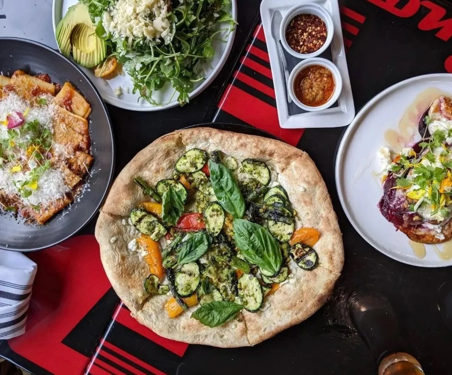 A table with various dishes including a pizza with basil and vegetables, a salad with greens, cheeses, and dressing, a plate of food with microgreens and possibly meat, and three small bowls of dipping sauces.