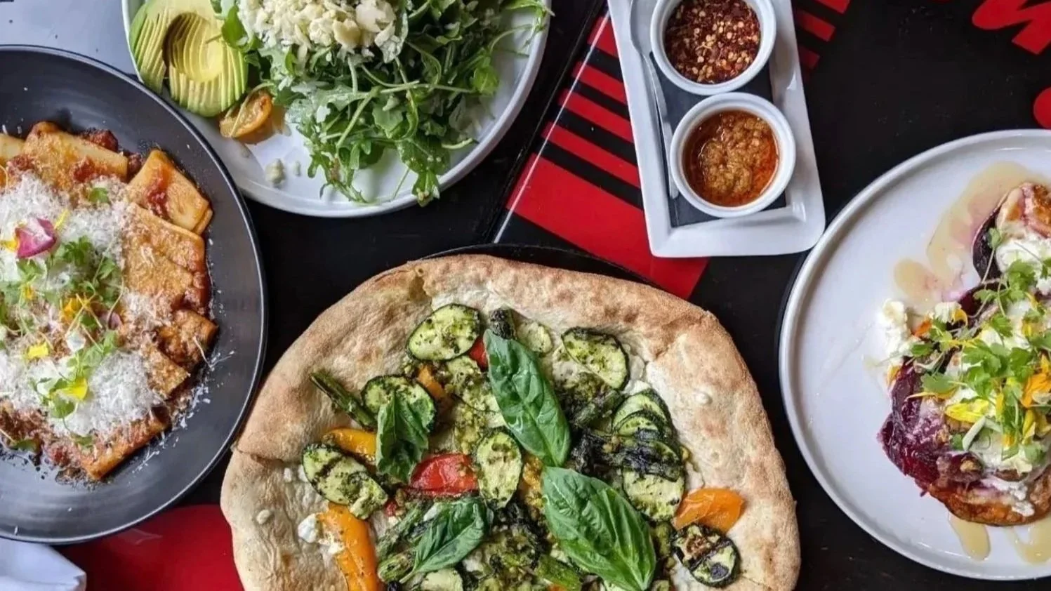 Assorted dishes including a pizza topped with zucchini and basil, a plate of salad with greens and avocado, a slice of lasagna garnished with cheese and herbs, and a dish topped with meat and sauce.