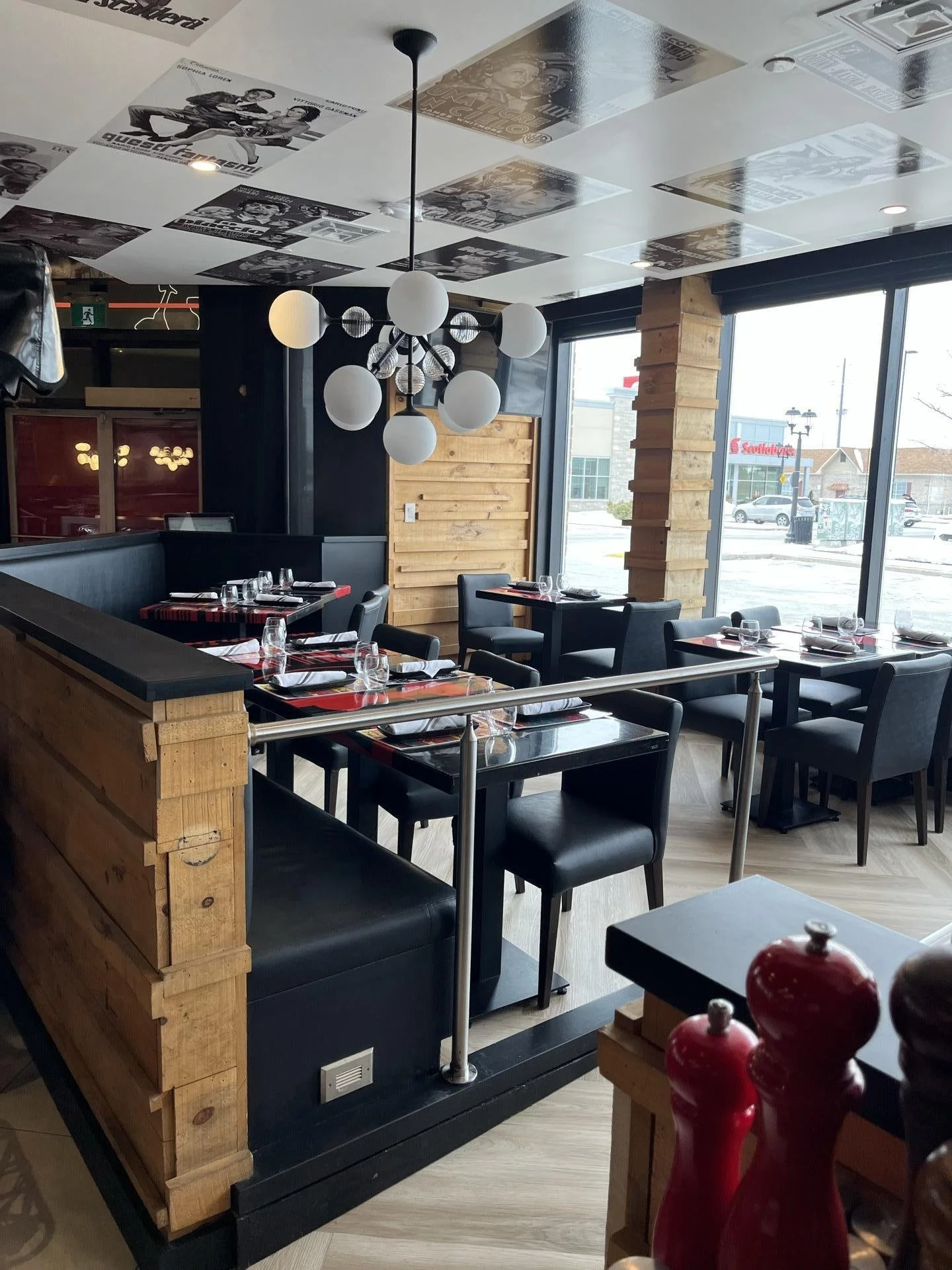Interior of a restaurant with black chairs, tables set with glasses and napkins, large windows, modern chandelier, black and wood decor, and ceiling with black-and-white photographs