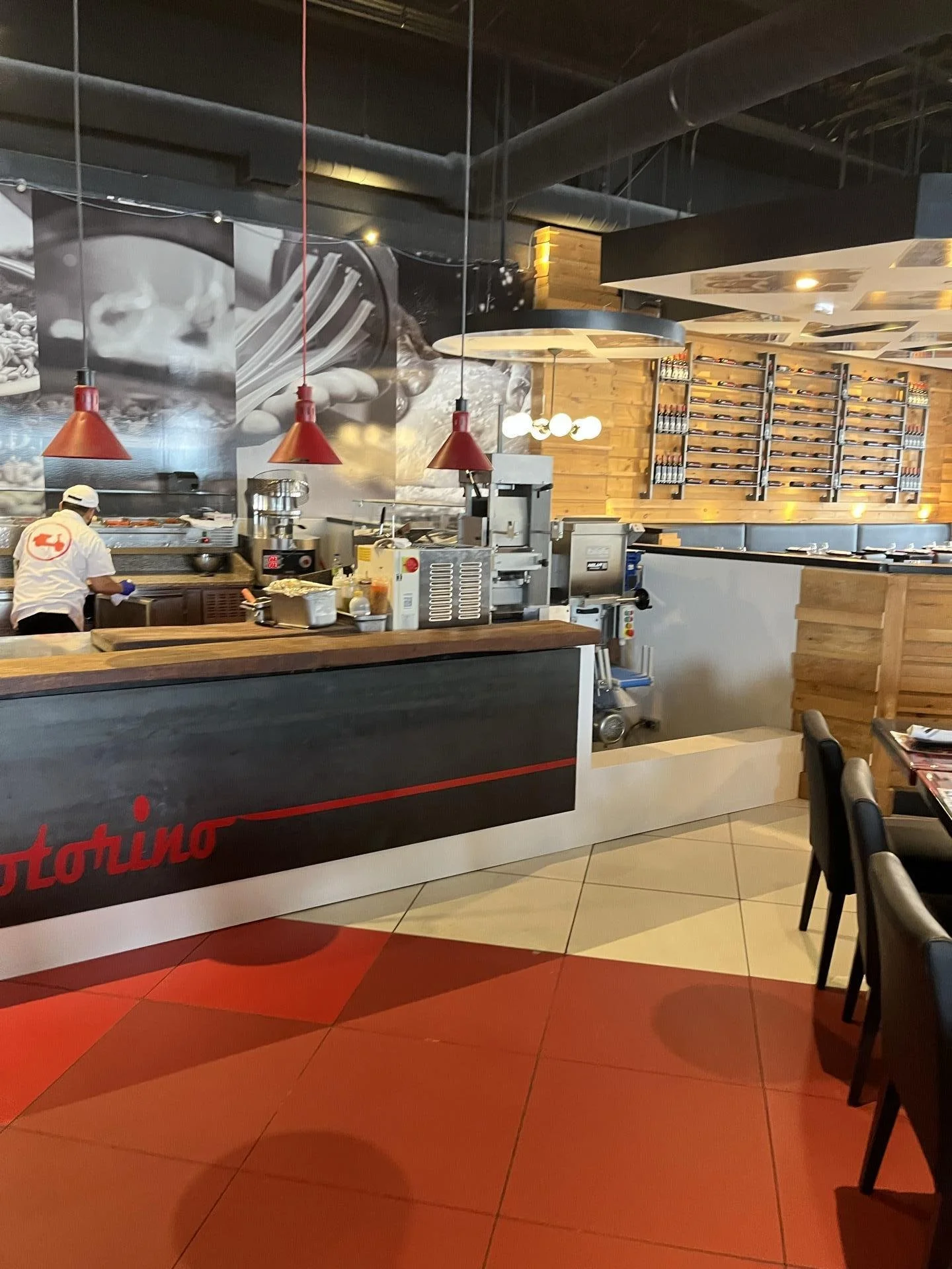 Inside a modern restaurant with a pizza kitchen, showing a chef preparing food, several red pendant lights, wood decor, and a display of wine bottles on the wall.